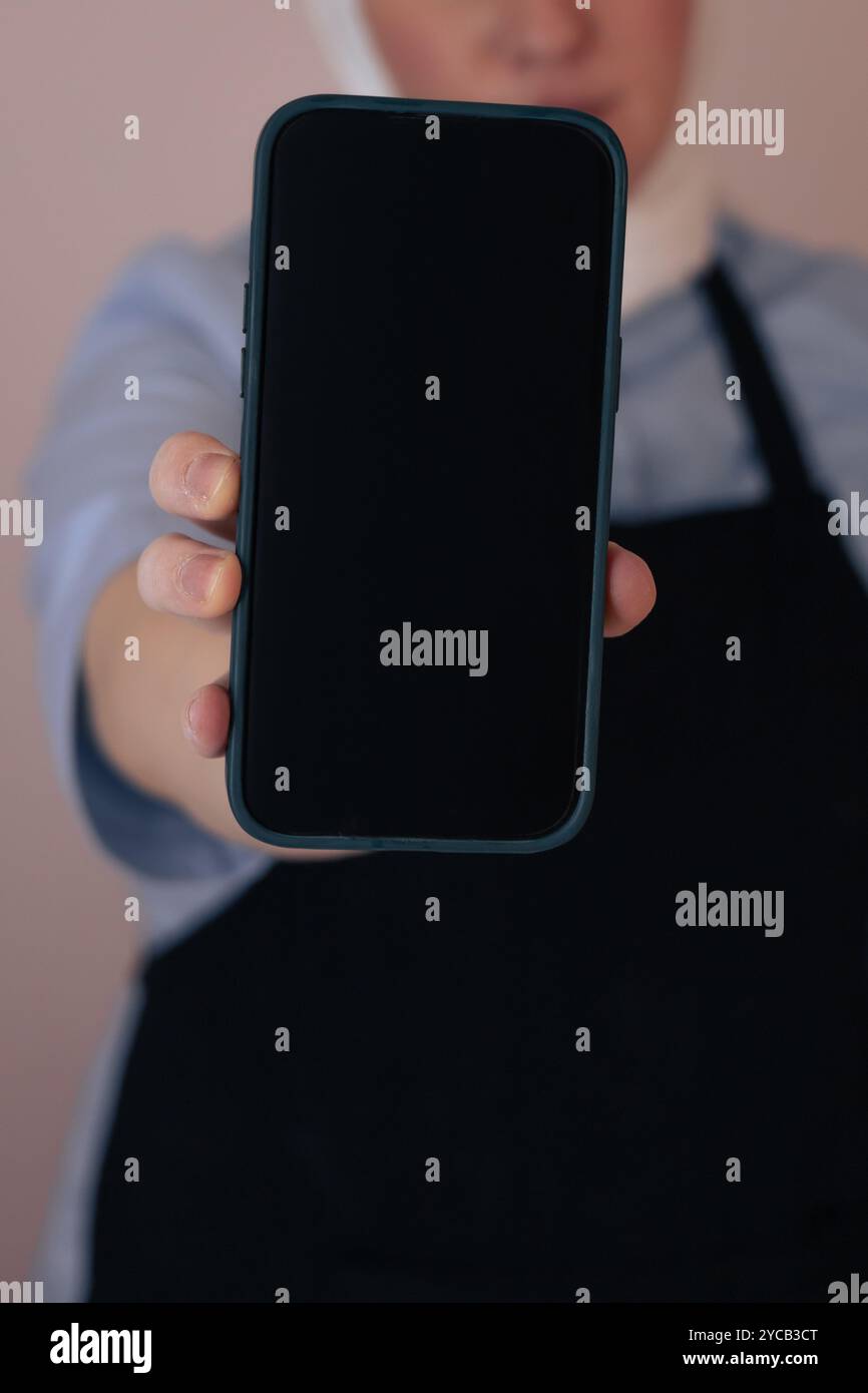 Eine Frau auf der Schürze hält ein leeres Smartphone, während sie in einem sanft beleuchteten Raum steht Stockfoto