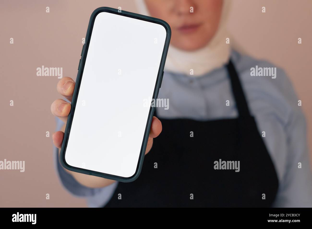 Eine Frau auf der Schürze hält ein leeres Smartphone, während sie in einem sanft beleuchteten Raum steht Stockfoto