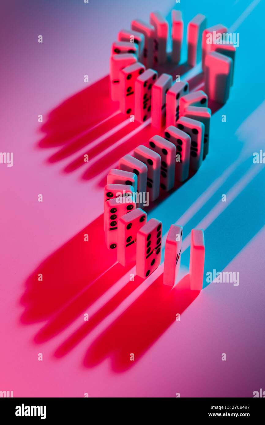 Eine kaskadierende Linie aus weißen Dominosteinen, die von Neonrot und Blau beleuchtet werden und eine Retro-Atmosphäre und Kindheitserinnerungen wecken. Stockfoto