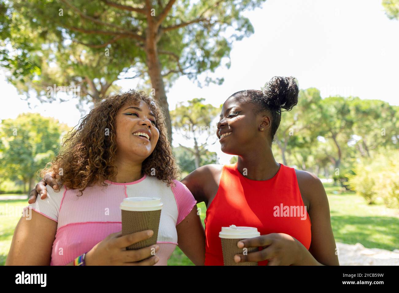 Zwei glückliche Freunde, ein Afroamerikaner und ein Lateinamerikaner, genießen einen sonnigen Tag im Park, teilen sich ein Lächeln und halten Kaffeetassen Stockfoto