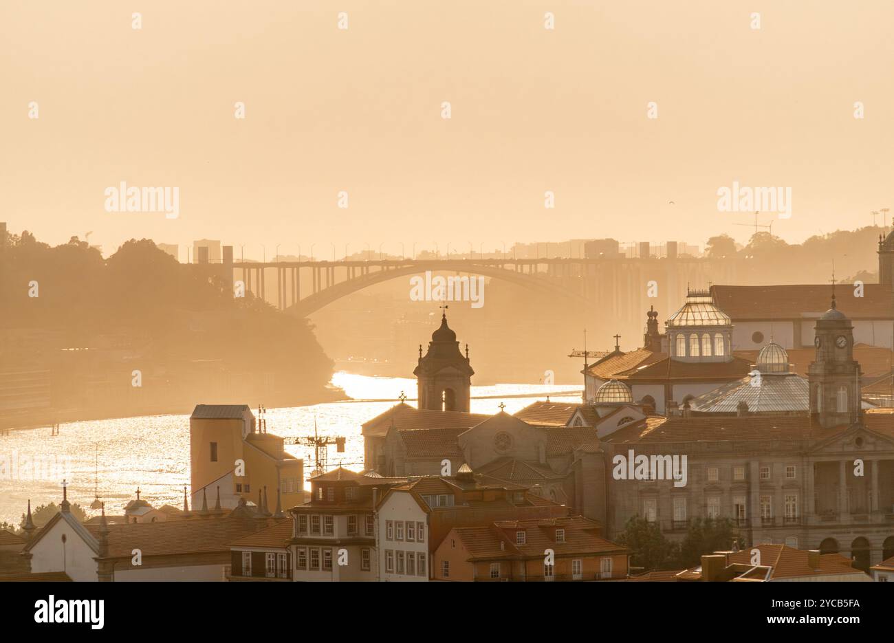 Die ruhige Szene von Porto bei Sonnenuntergang mit goldenem Glanz über historischen Gebäuden und dem Fluss Douro, Einer Silhouette einer entfernten Brücke Stockfoto