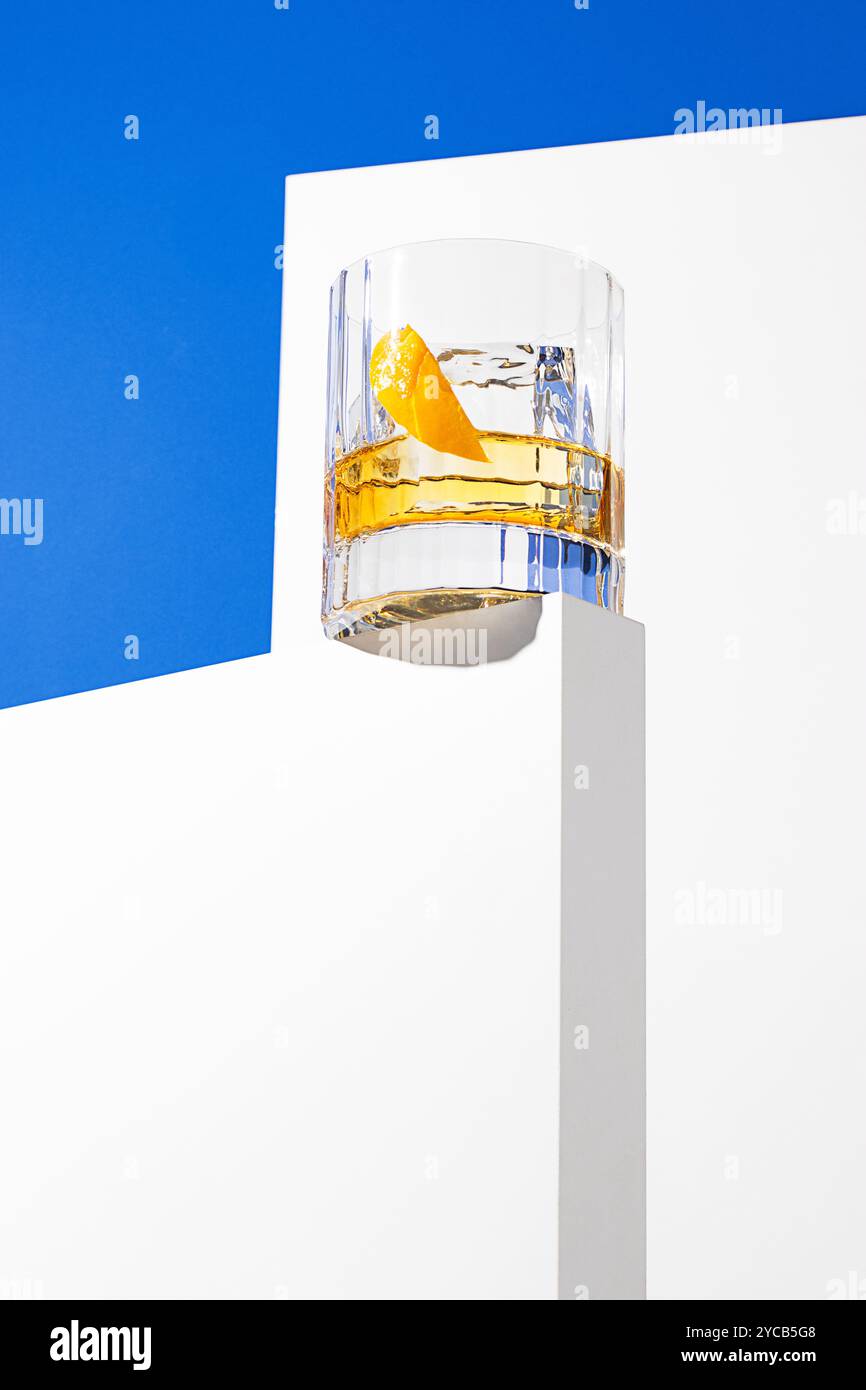 Minimalistischer Schuss eines Whiskyglases mit Orangenschalen auf geometrischem weißem und blauem Hintergrund Stockfoto
