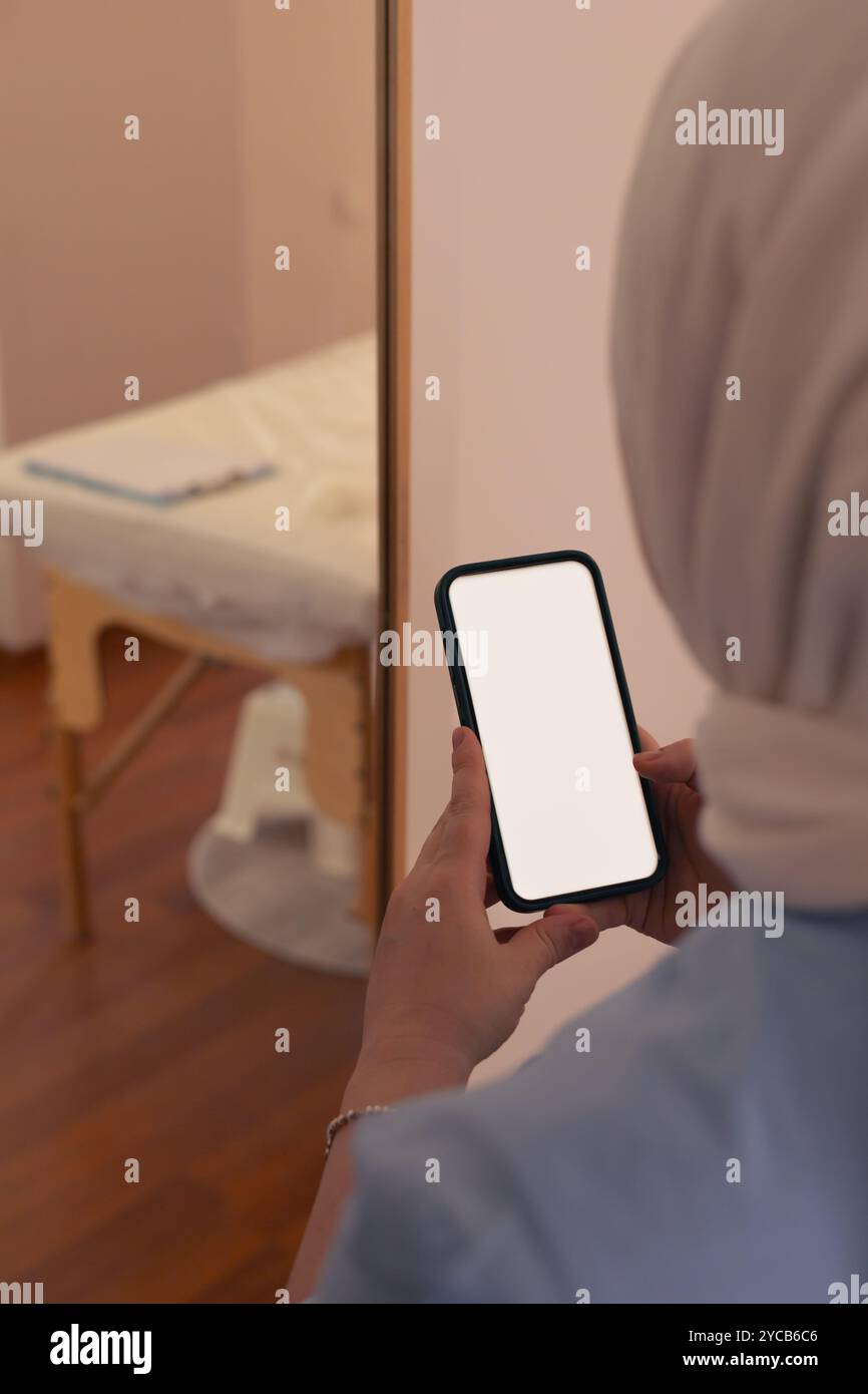 Eine Frau auf der Schürze hält ein leeres Smartphone, während sie in einem sanft beleuchteten Raum steht Stockfoto