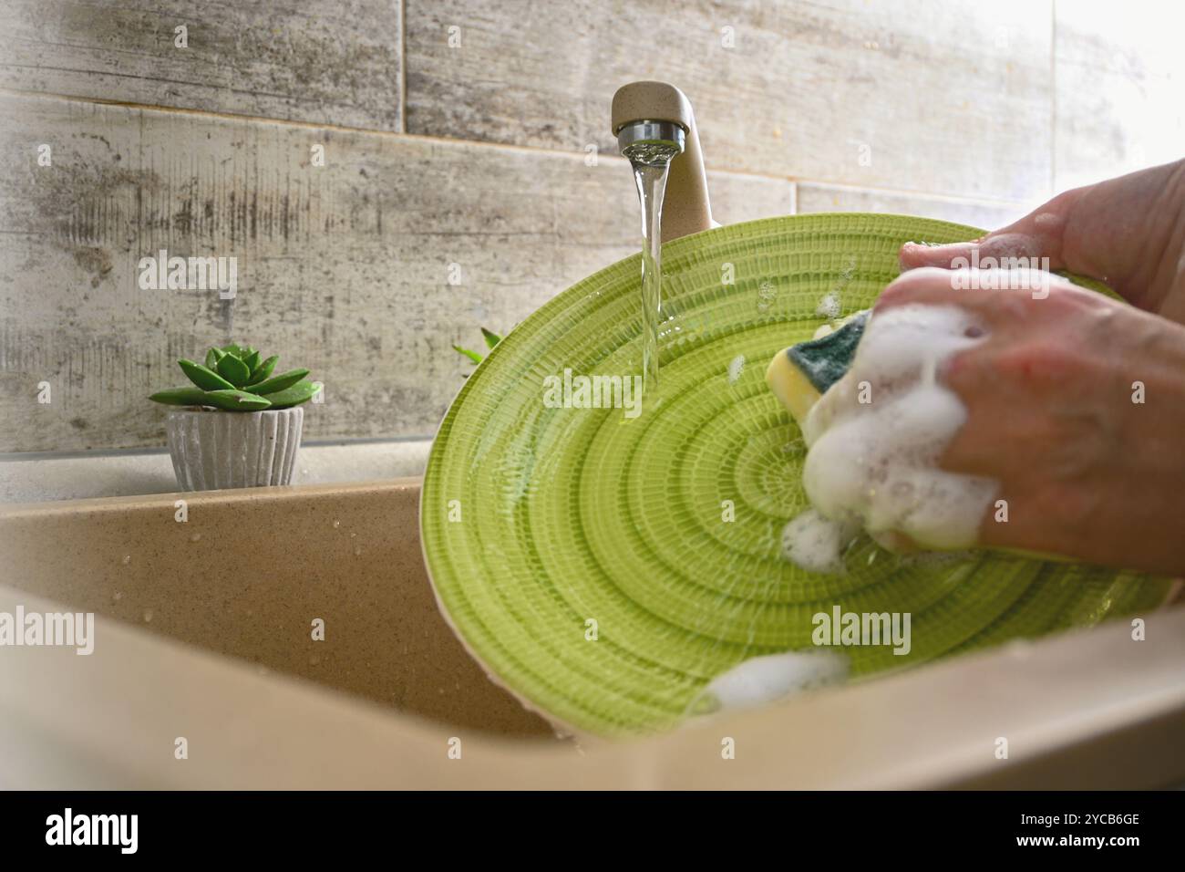 Hände waschen einen grünen Teller mit einem gelben Schwamm unter fließendem Wasser in einem Spülbecken. Stockfoto