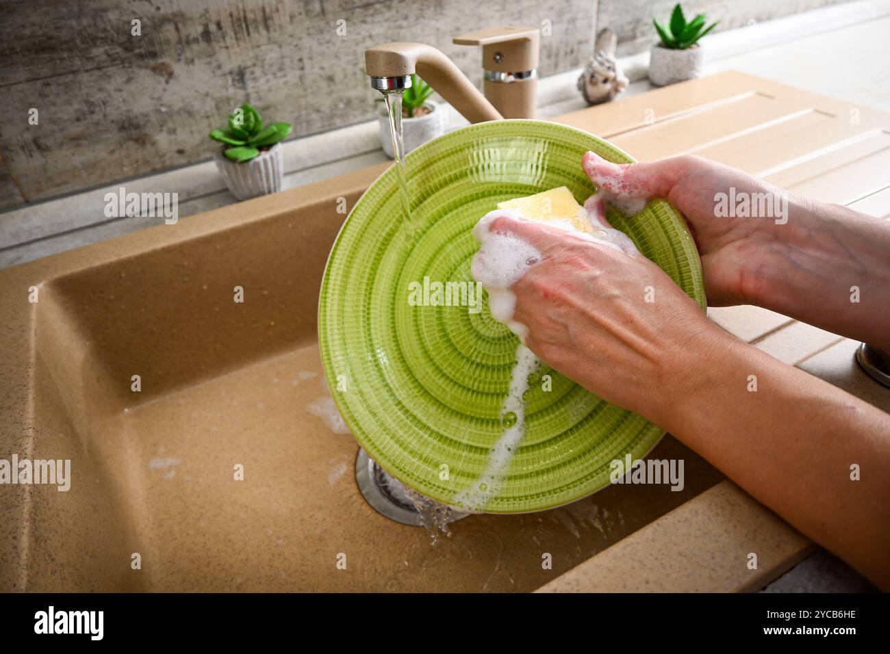 Hände waschen einen grünen Teller mit einem gelben Schwamm unter fließendem Wasser in einem Spülbecken. Stockfoto