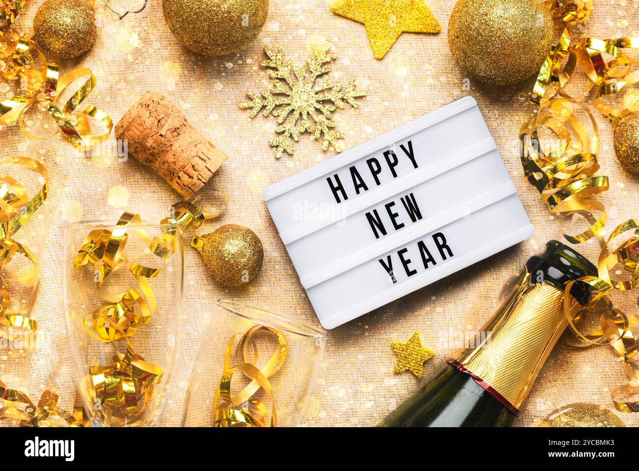 Happy New Year 2025. Top-Blick auf Leuchtkasten mit dem Wort Happy New Year 2025, Kristallgläser und goldene weihnachtskugeln auf einer goldenen Glitzertischdecke. Ne Stockfoto