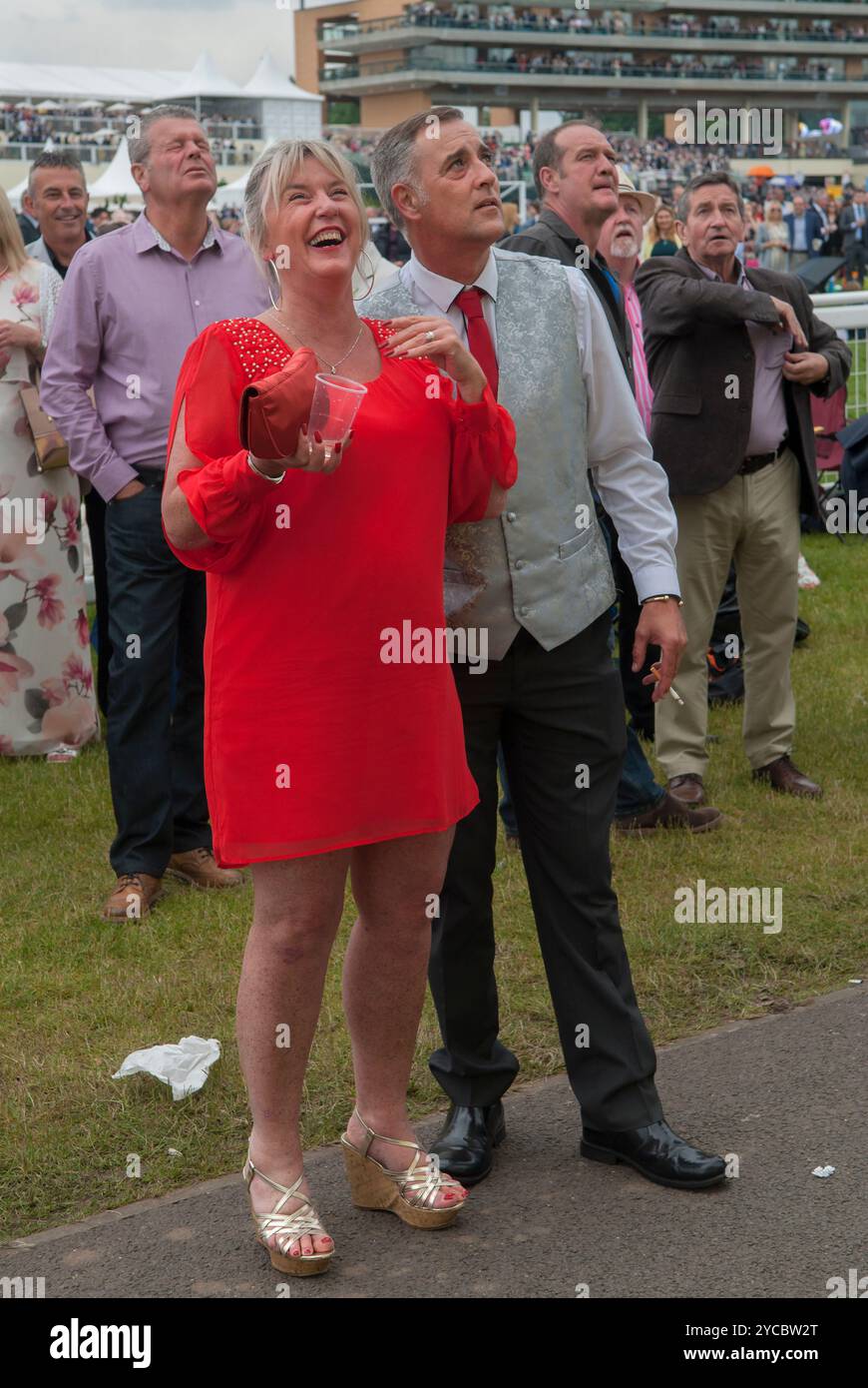 Fröhlich, lachend, lächelnd eine Frau mit ihrem Partner, die das Pferd auf einer großen Außenleinwand beobachtet. Sie sind auf der Heide, der freien Seite der Rennbahn. Ich würde sagen, ihr Pferd gewinnt, ihr Ehemann sieht nicht so glücklich aus. Kontrastierender Look auf ihren Gesichtern. Ascot, Berkshire, England 18. Juni 2016 2010, UK HOMER SYKES Stockfoto