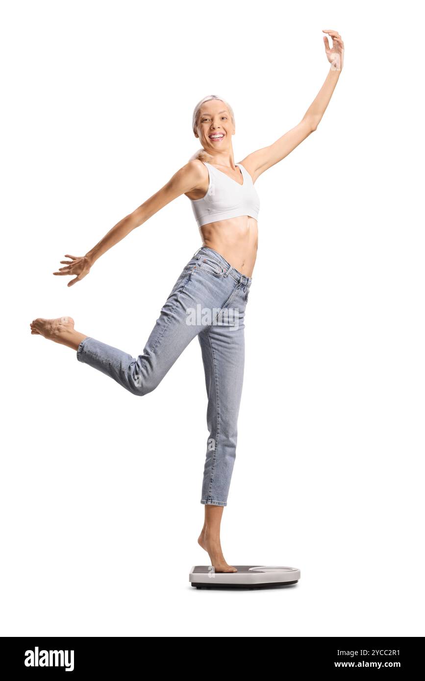 Große schlanke Frau in Jeans und weißem Crop Top, die auf einer Waage auf weißem Hintergrund tanzt Stockfoto