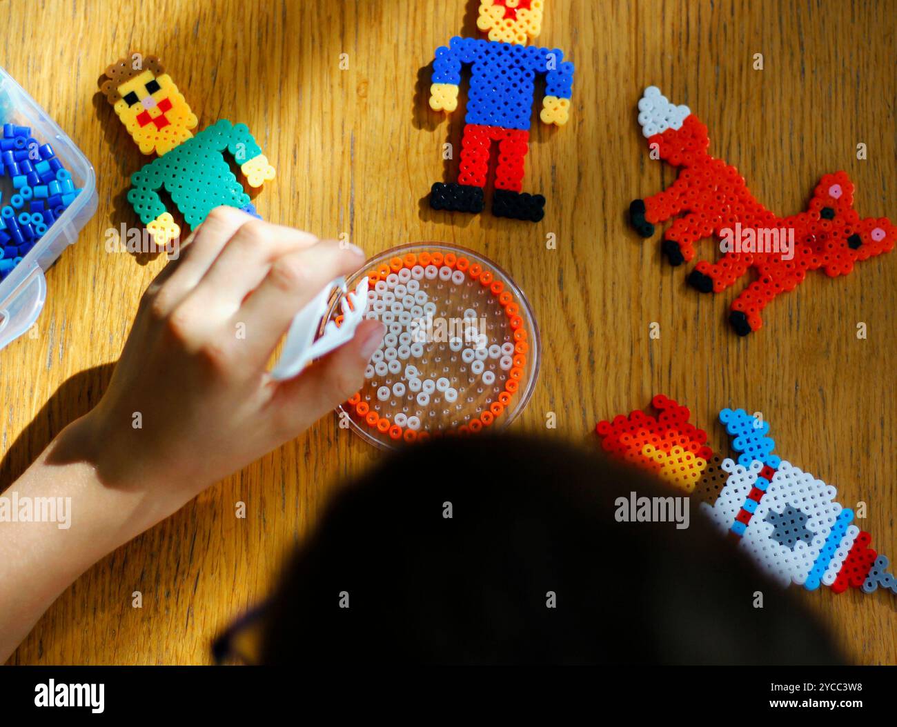 Die Hand hält eine Pinzette mit einer Perle, um Kunstspielzeug aus schmelzbaren bunten Perlen (auch Perlperlen oder Eisenkegeln) zu kreieren. Spielzeugentwickelnde ima für Kinder Stockfoto