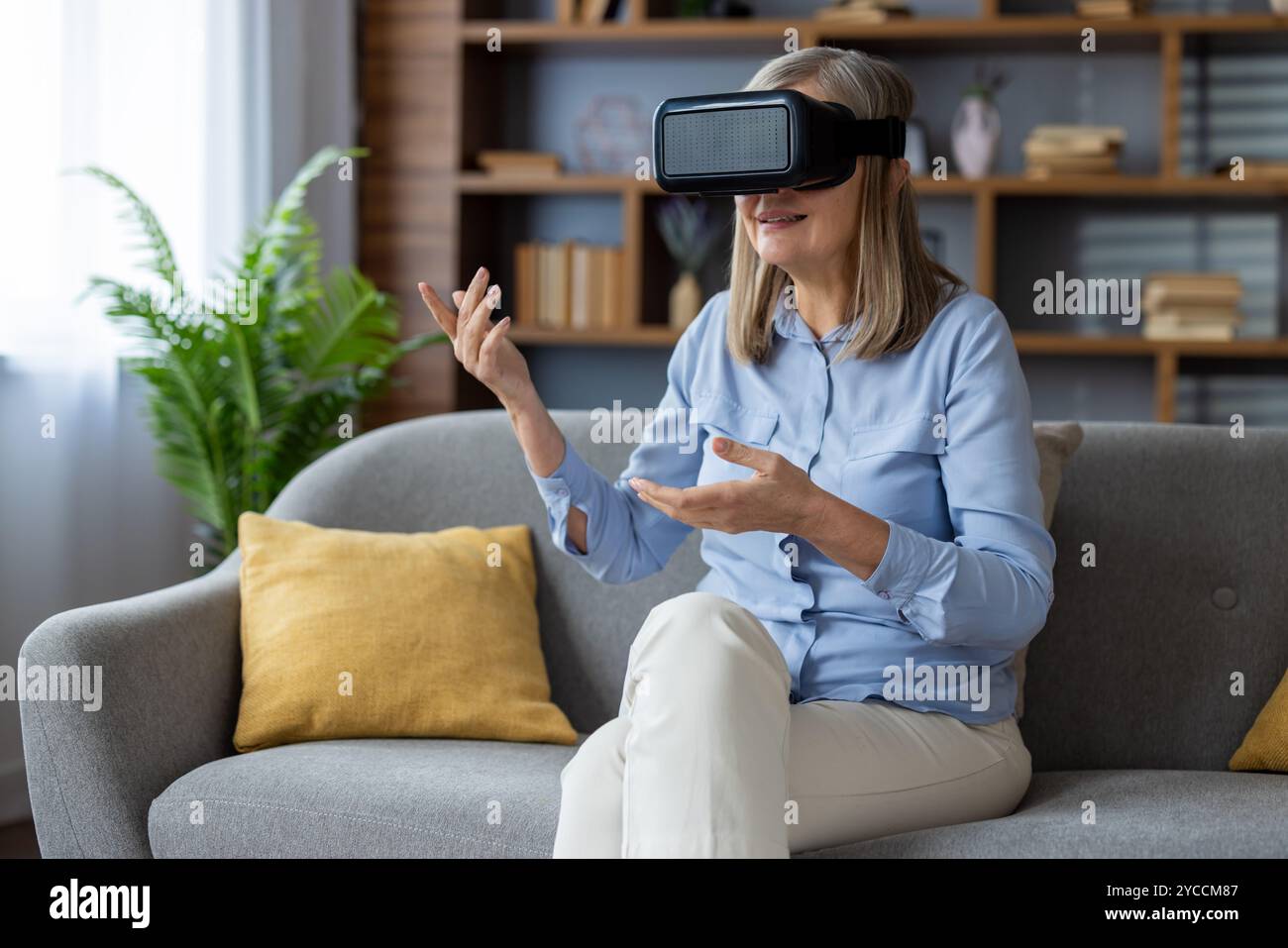 Die Seniorin verwendet das VR-Headset auf dem grauen Sofa und erlebt die immersive Virtual-Reality-Technologie, drückt Begeisterung aus, drückt Gesten mit den Händen aus, Enjo Stockfoto