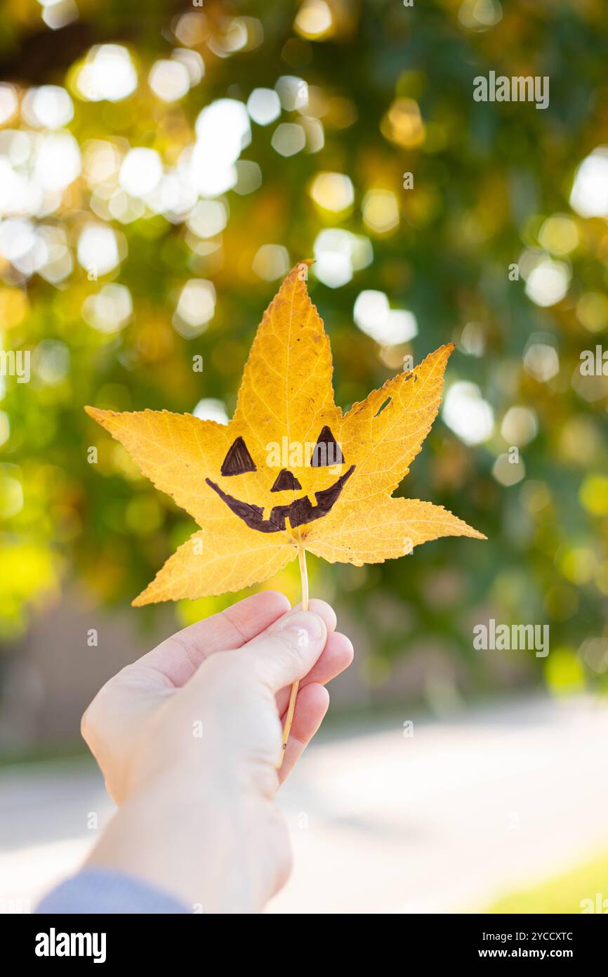 Die Hand hält ein gelbes flüssiges Blatt mit der Zeichnung eines halloween Kürbisgesichts drauf. Herbsthintergrund mit Kopierraum. Vertikale Aufnahme. Stockfoto