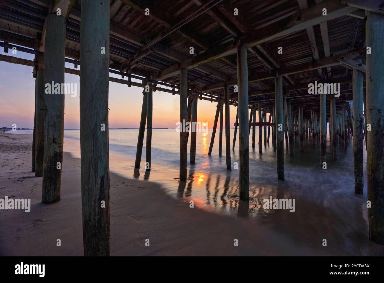 Sonnenlicht filtert durch die hölzernen Stützen eines Piers und wirft Reflexionen auf den nassen Sand darunter. Die ruhige Meereskulisse verstärkt den friedlichen geldautomaten Stockfoto