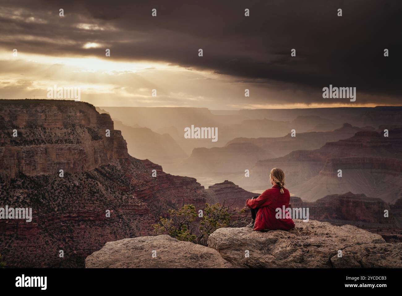 Eine Person sitzt kontemplativ auf einem felsigen Felsvorsprung mit Blick auf den riesigen und beeindruckenden Grand Canyon. Der Himmel ist voller dramatischer Wolken wie die Sonne. Stockfoto