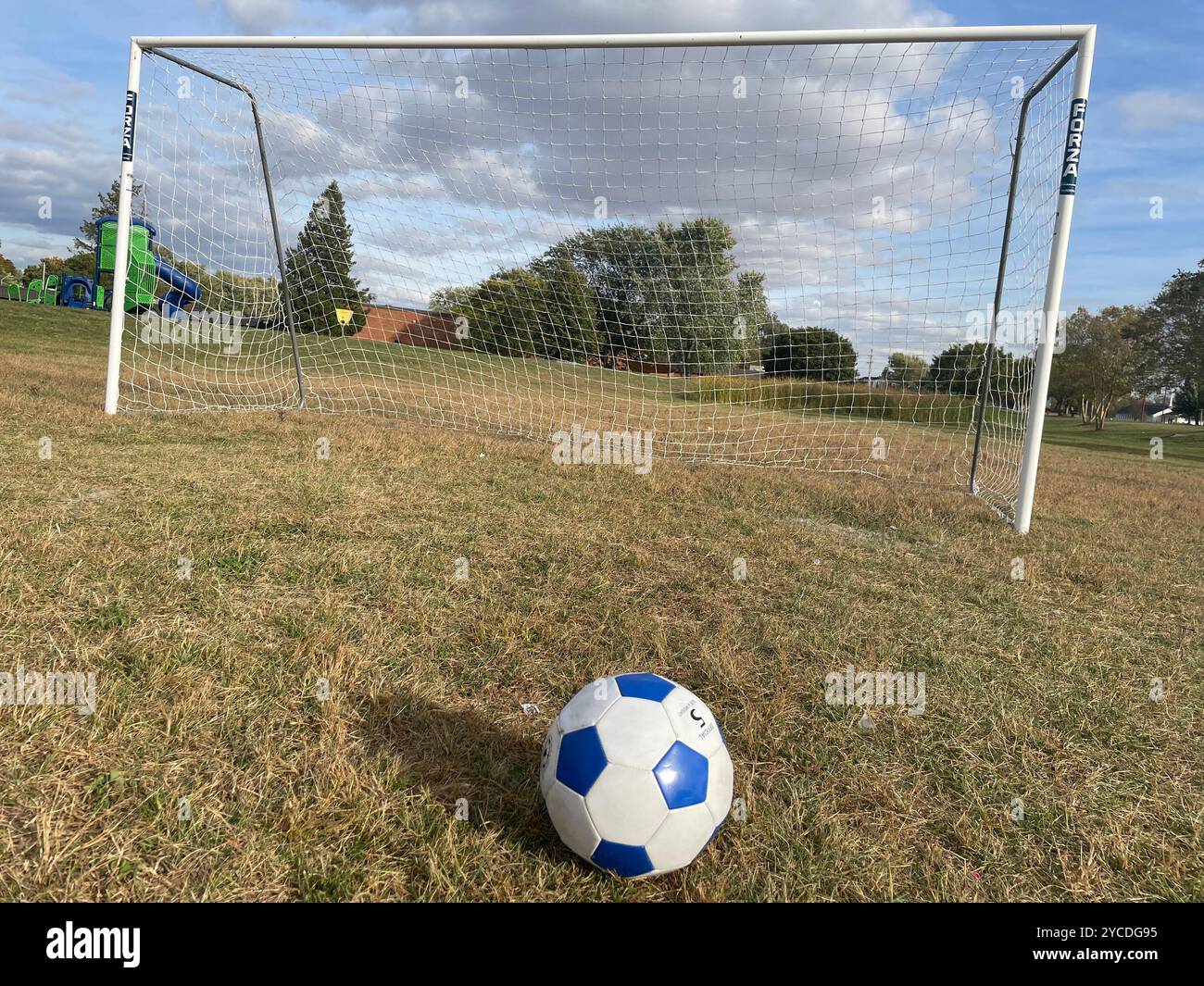 Fußball, der vor einem Tor auf einem trockenen, grasbewachsenen Feld unter teilweise bewölktem Himmel steht, symbolisiert Spiel, Sport und Erholung im Freien. - Smartphone-aufgenommenes Stockfoto