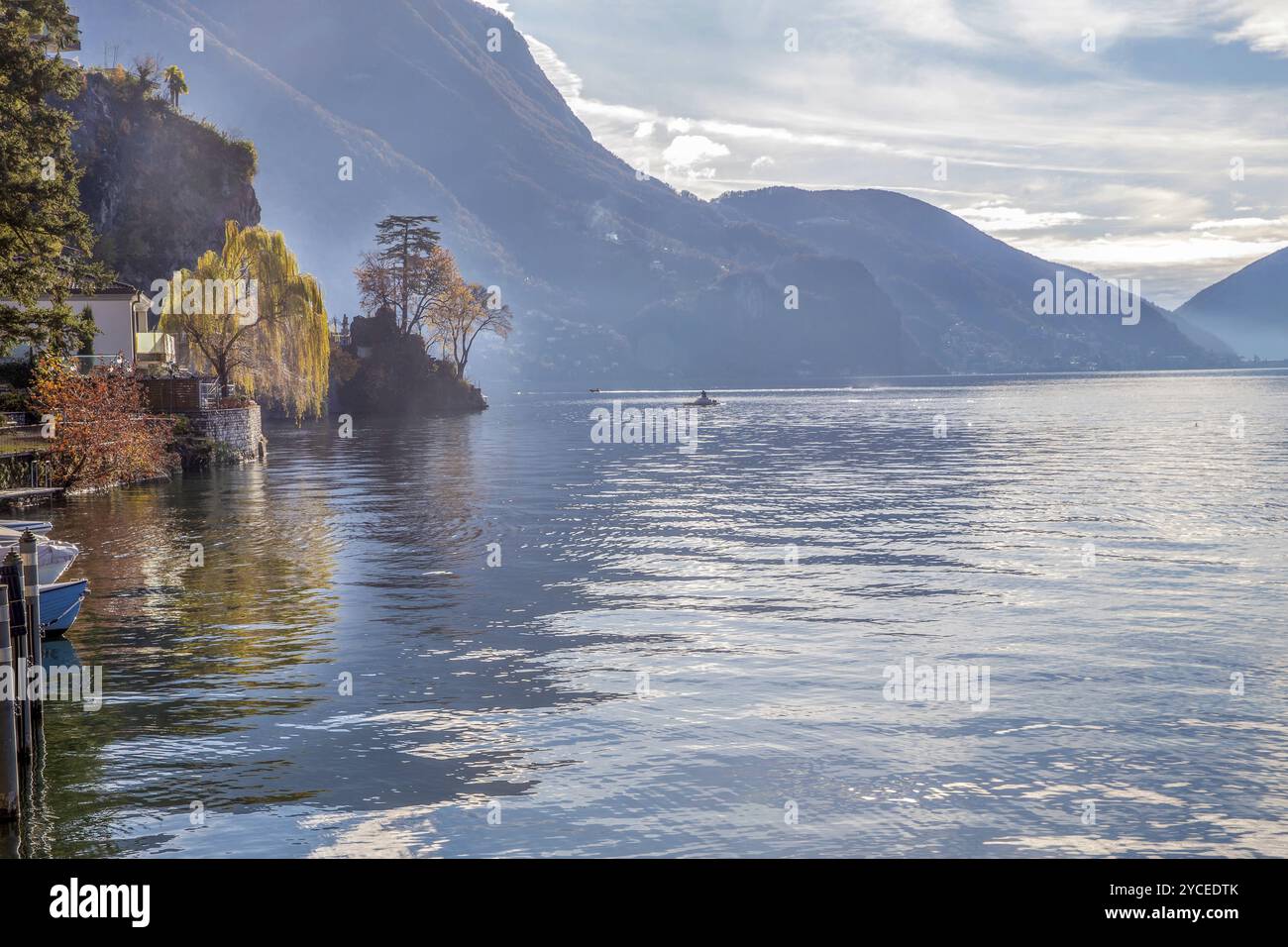 Herrlicher Blick auf den Luganer See mit blattfarbenen Herbstbäumen und Booten im Wasser. Ein Schnellboot fährt in der Ferne und ein festgemachtes Boot fährt Kajaks (jetzt Stockfoto