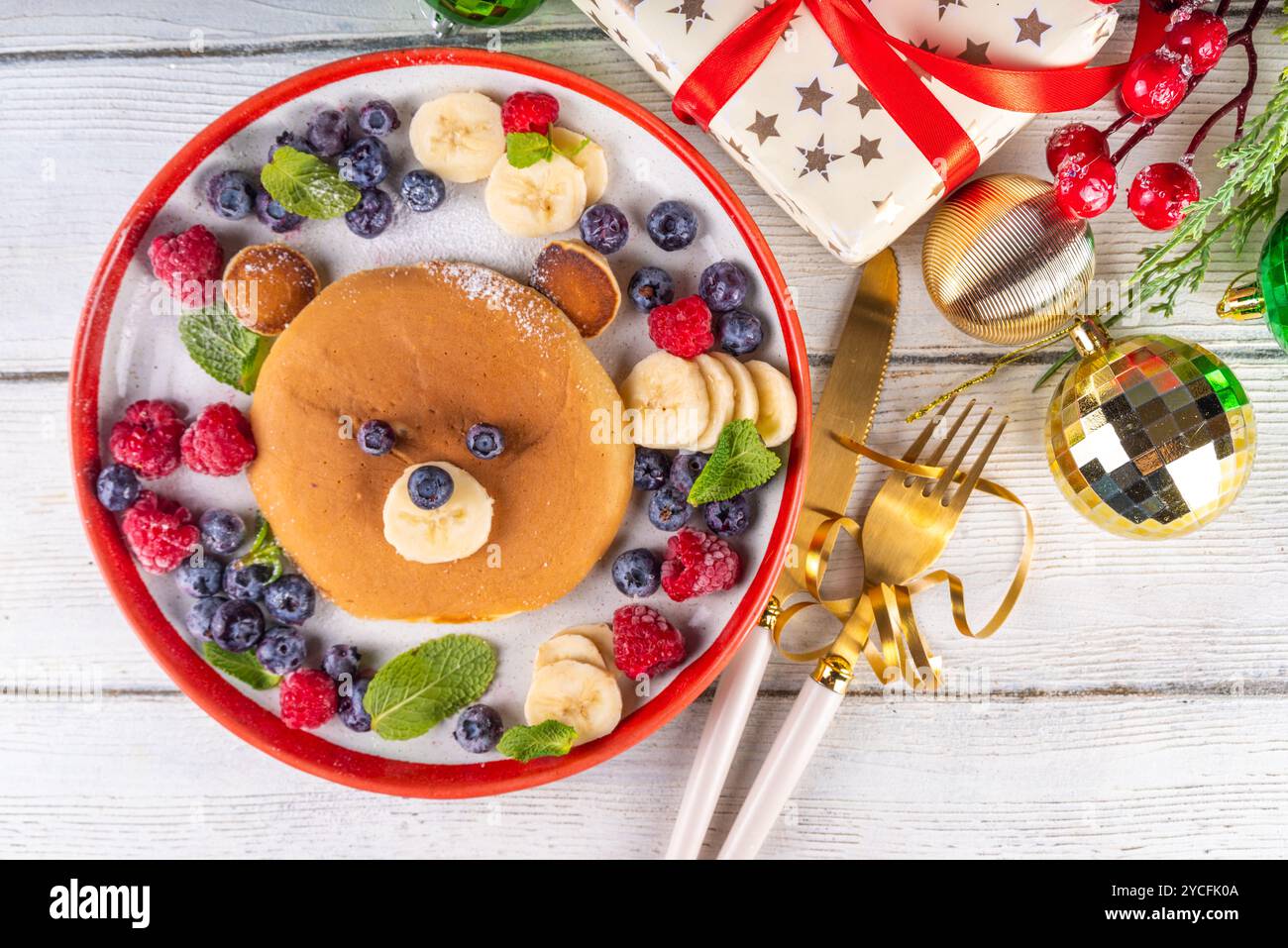 Lustige und süße Weihnachts-Teddybären-Pfannkuchen auf einem rot-weißen Teller, dekoriert mit frischen Beeren, Obst und Puderzucker, Kinderweihnachtsfrühstück Stockfoto