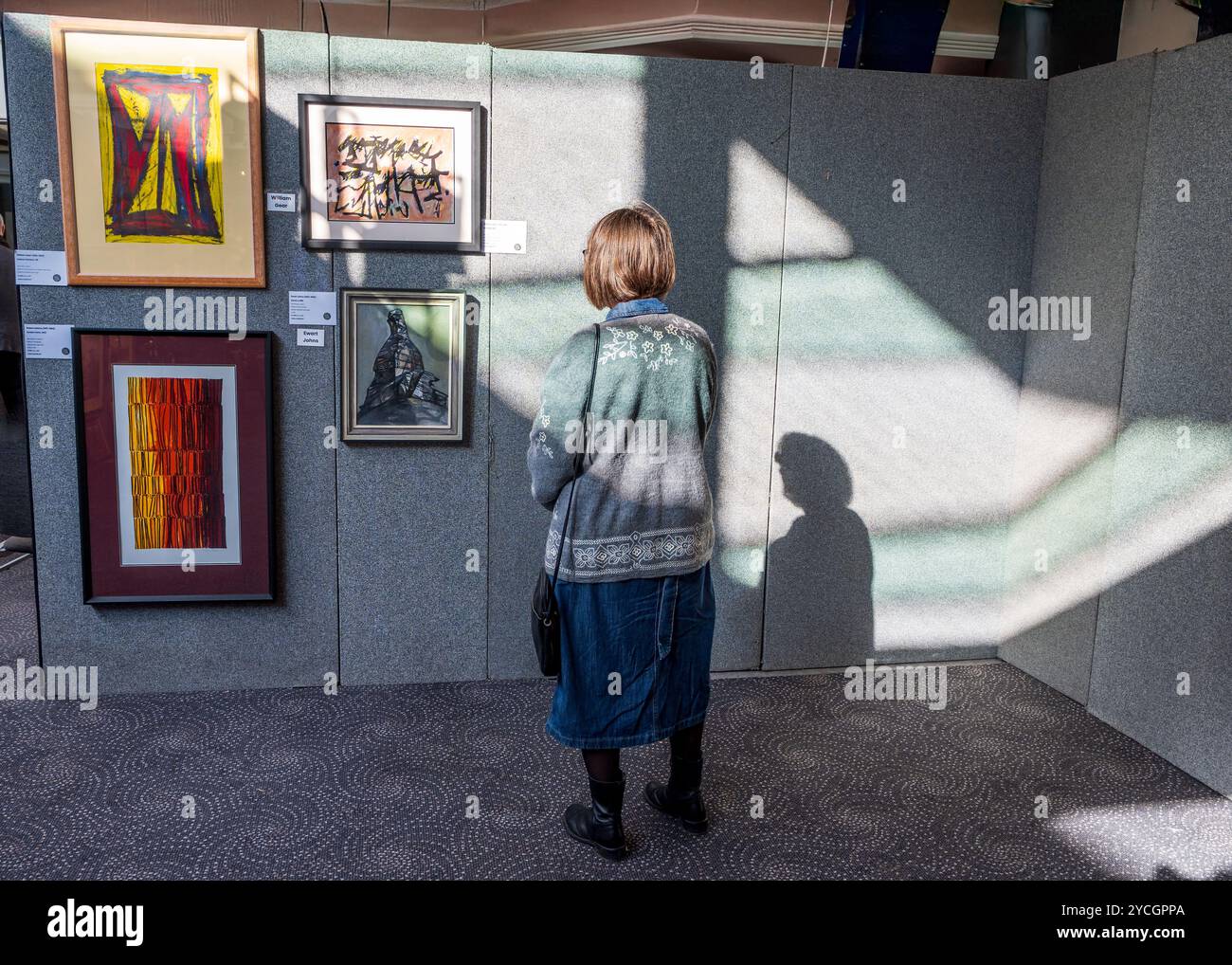 Frau, die Gemälde an einer sonnendurchfluteten Galeriewand studiert, wirft ihren Schatten neben den Bildern. Stockfoto