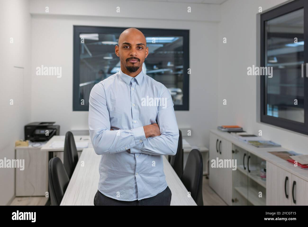 Selbstbewusster junger afroamerikanischer Mann, stolz posiert in einem Büro mit gekreuzten Armen, Porträt aufgenommen. Professionalität und Expertise in Unternehmenskonzepten Stockfoto