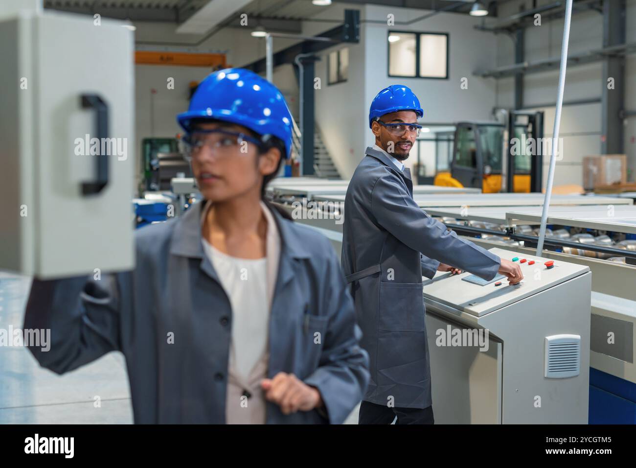 Zwei Fabrikingenieure, eine Inderin und ein afroamerikanischer Mann, überwachen Schalttafeln und den automatisierten Fertigungsprozess. Stockfoto