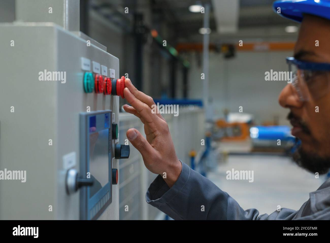 Afroamerikanischer Ingenieur, der einen grauen Arbeitsmantel und einen blauen Helm trägt und die Produktion und Programmierung einer Maschine in einer Fabrik steuert. Stockfoto