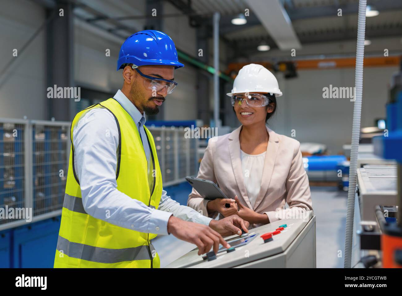 Die indische Betriebsleiterin trainiert einen afroamerikanischen männlichen Mitarbeiter in einem blauen Helm und einer gelben Weste, um Maschinen zu bedienen. Stockfoto
