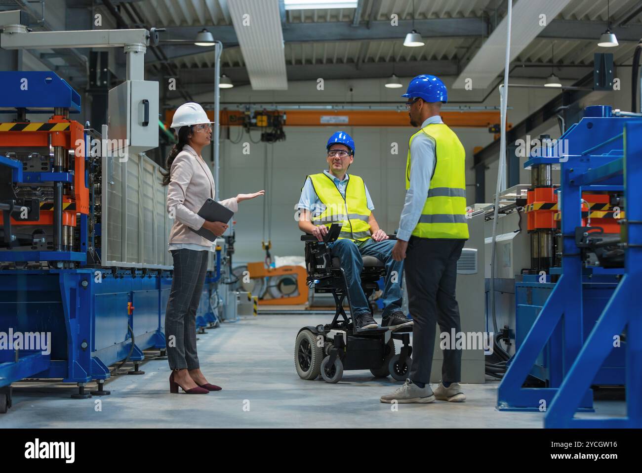 Weibliche Führungskraft spricht mit zwei Arbeitern an der Produktionslinie, darunter einer mit Behinderung, über den Produktionsprozess. Stockfoto