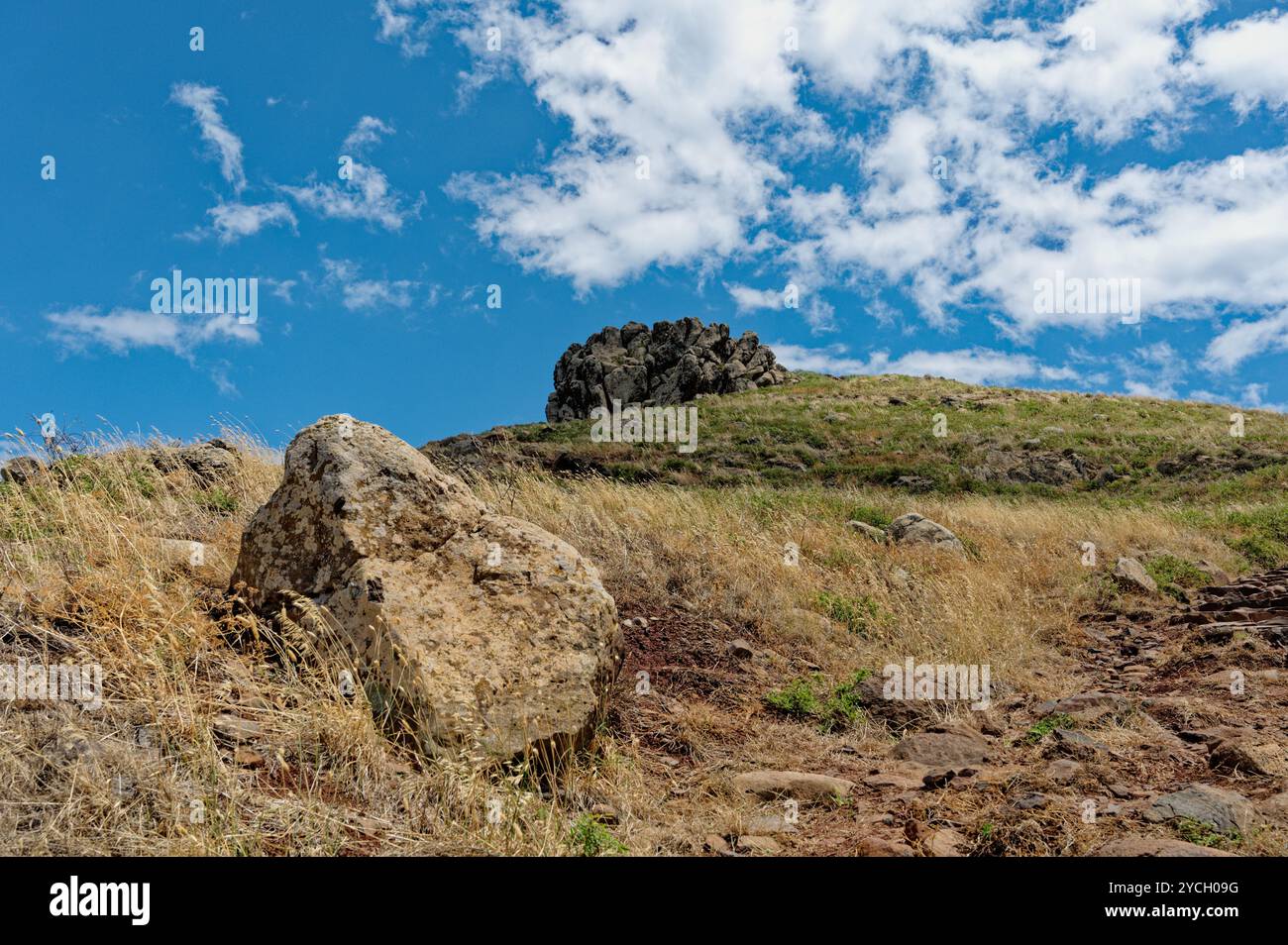 Ein großer Felsbrocken liegt auf einem grasbewachsenen Hügel unter einem hellblauen Himmel mit einer zerklüfteten Felsformation in der Ferne bei Vereda da Ponta de São Lourenco Stockfoto