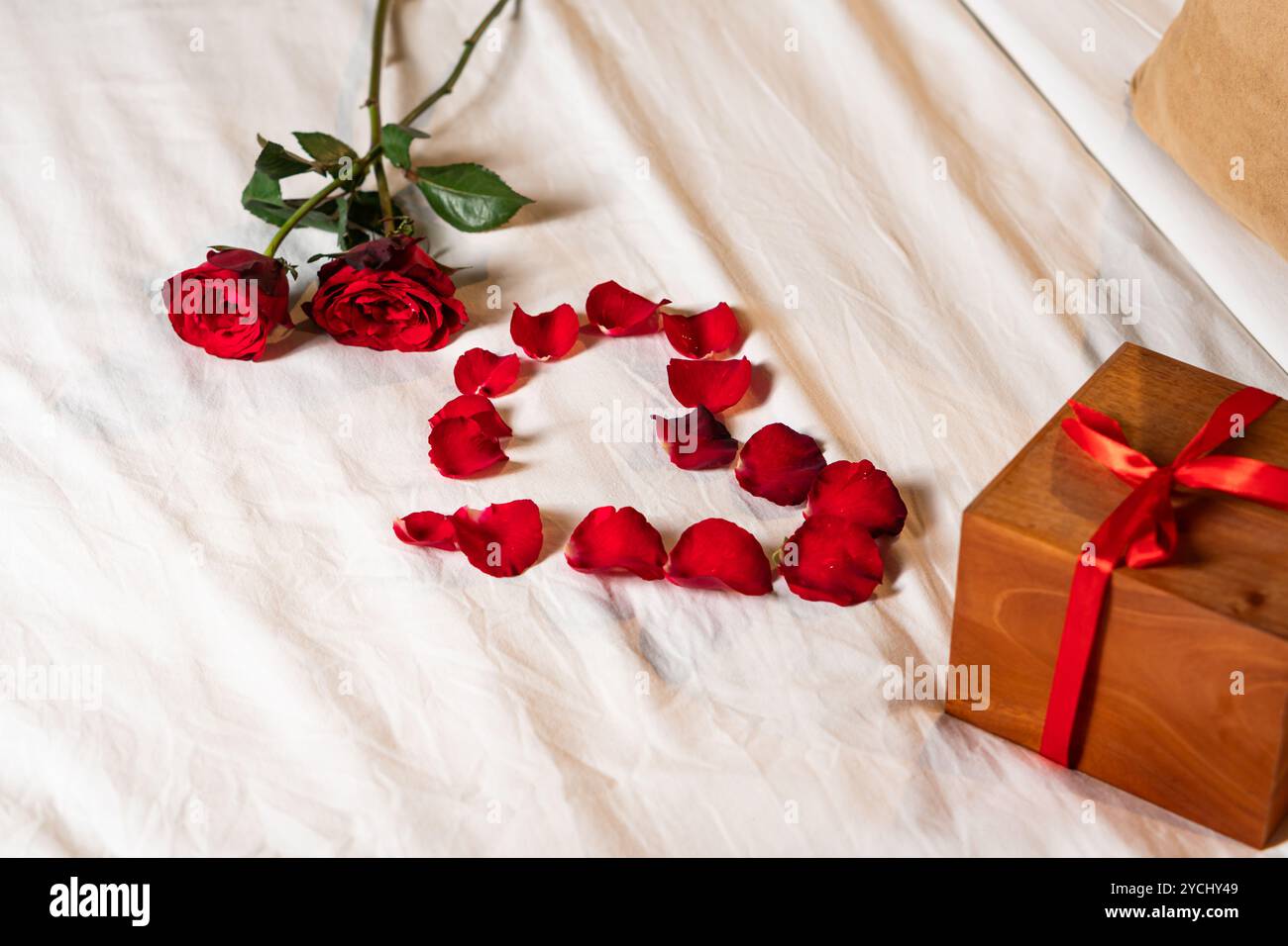 Romantische Einrichtung mit zwei roten Rosen und einer Herzform aus Rosenblättern auf weißem Bettlaken. Eine hölzerne Geschenkbox mit einem roten Band wird nea platziert Stockfoto