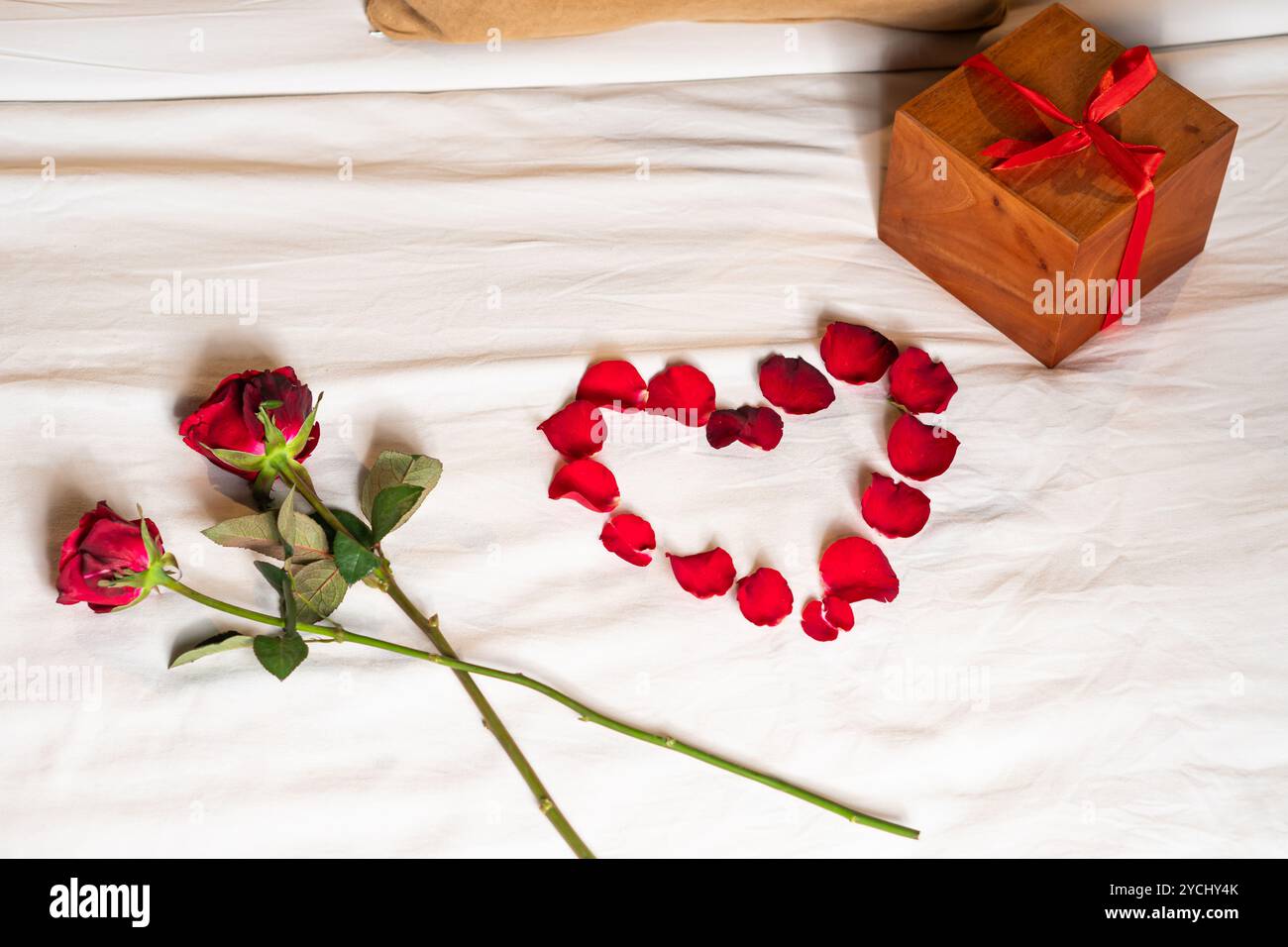 Romantische Einrichtung mit zwei roten Rosen und einer Herzform aus Rosenblättern auf weißem Bettlaken. Eine hölzerne Geschenkbox mit einem roten Band wird nea platziert Stockfoto