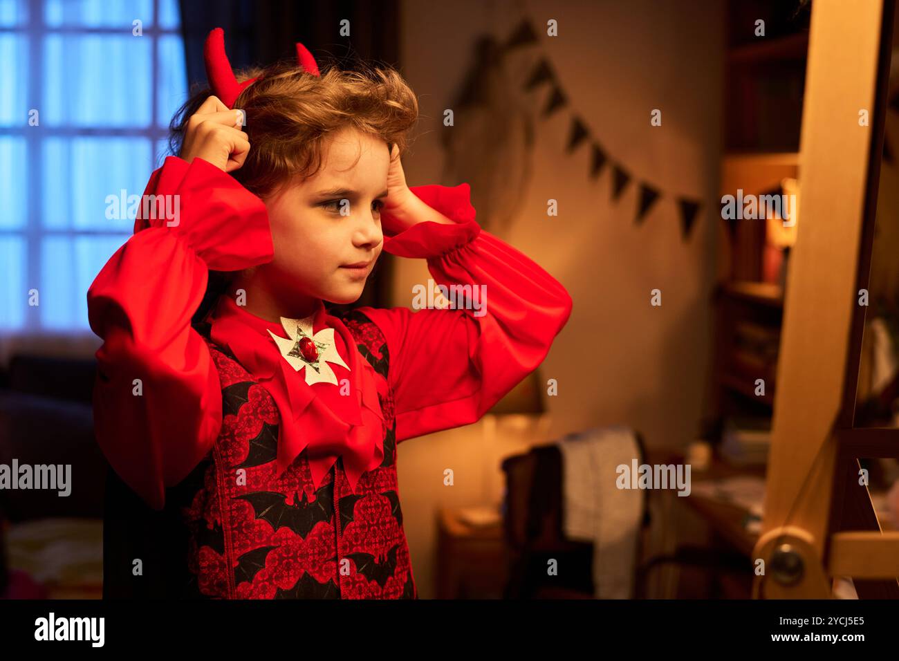 Niedlicher kleiner Junge im Teufelskostüm, der am Halloween-Abend vor dem Spiegel steht und ein Stirnband mit roten Hörnern anzieht Stockfoto