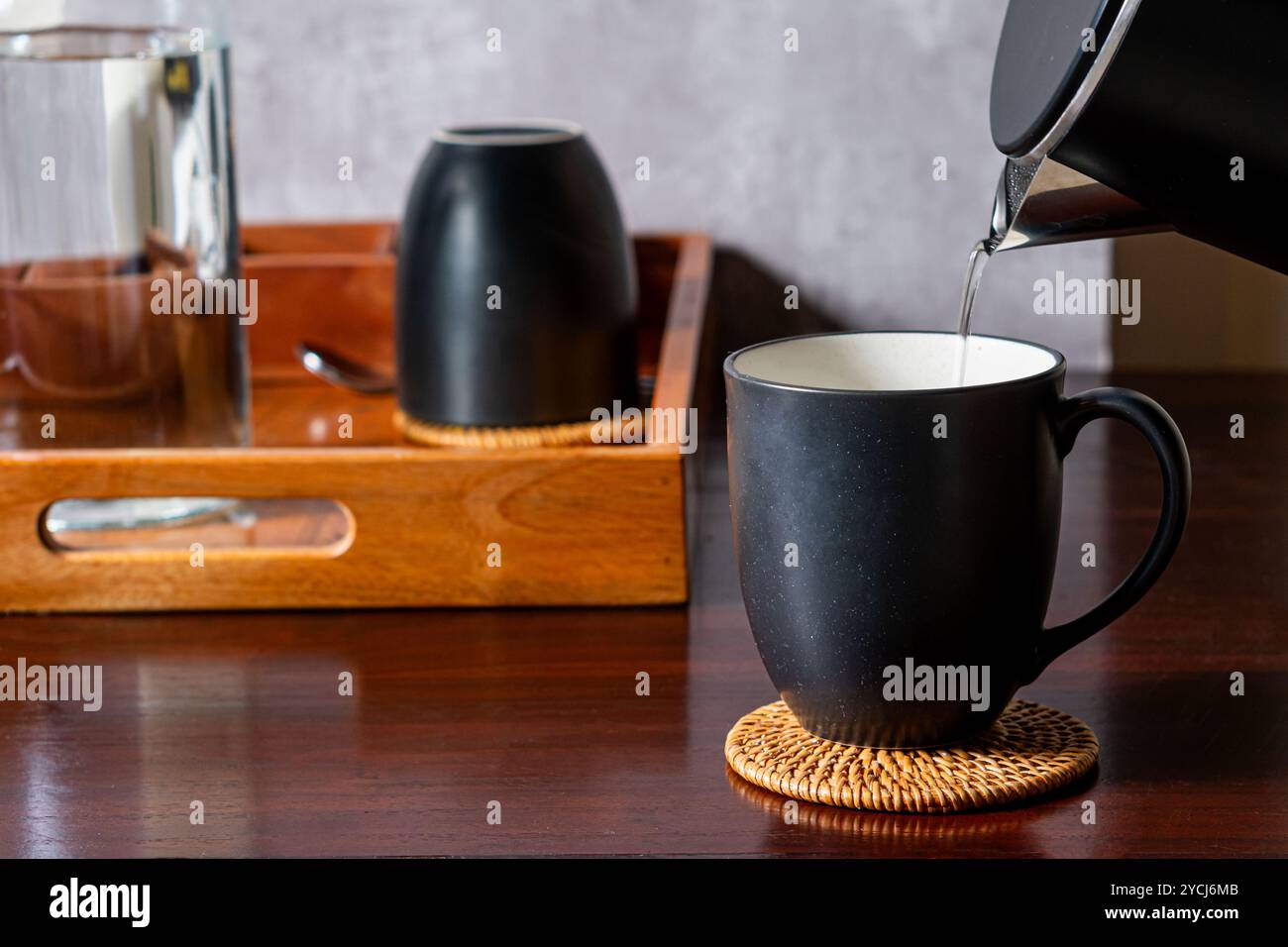 Eine schwarze Kaffeetasse auf einer gewebten Untersetzer, in der heißes Wasser aus einem Wasserkocher gegossen wird. Im Hintergrund befindet sich ein Holztablett mit einem Glas und einem Stockfoto
