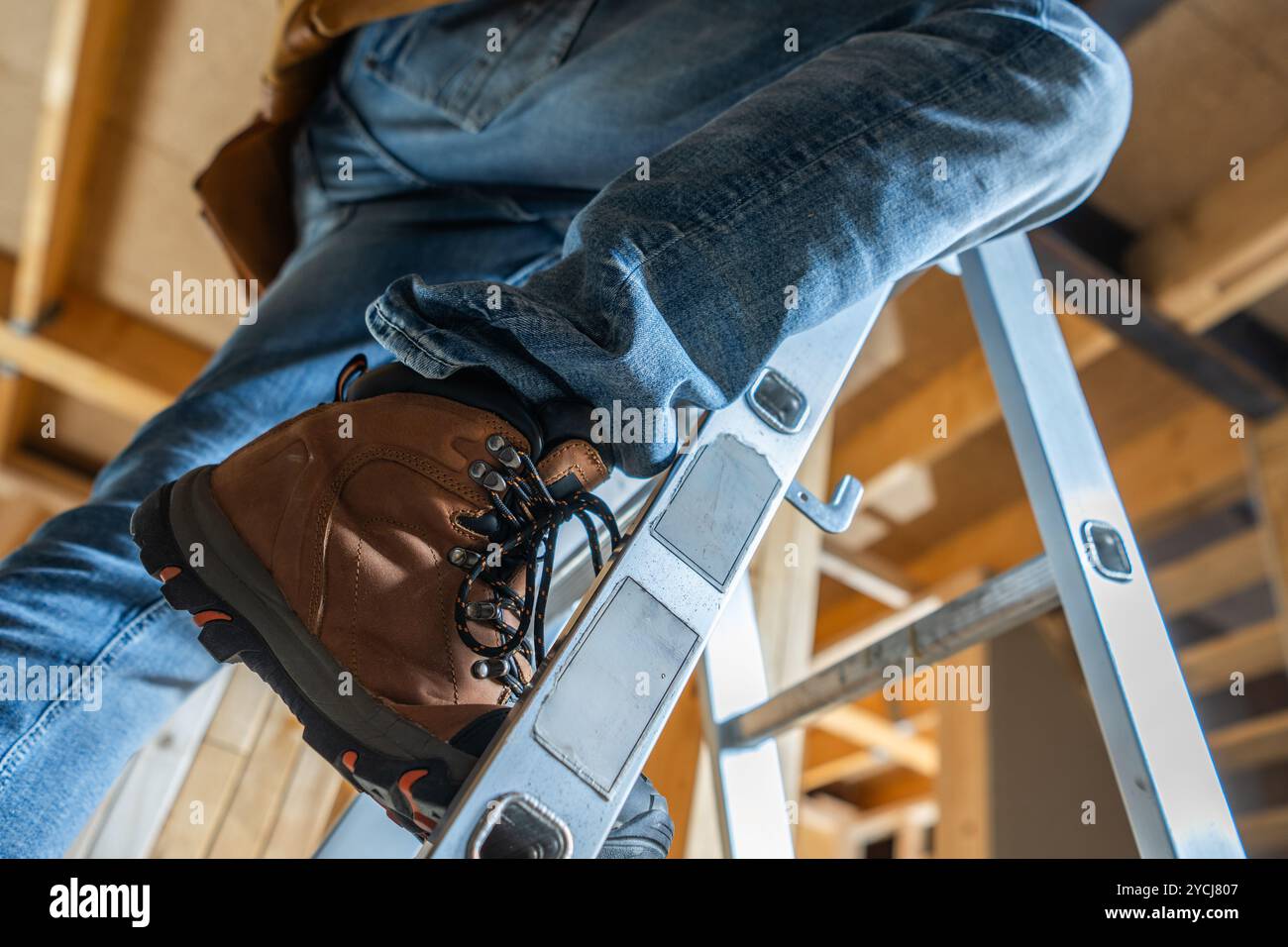 Ein Arbeiter wird sorgfältig auf einer Leiter positioniert, trägt stabile Stiefel und Jeans und arbeitet mit Renovierungsarbeiten unter hellen Deckenleuchten. Stockfoto
