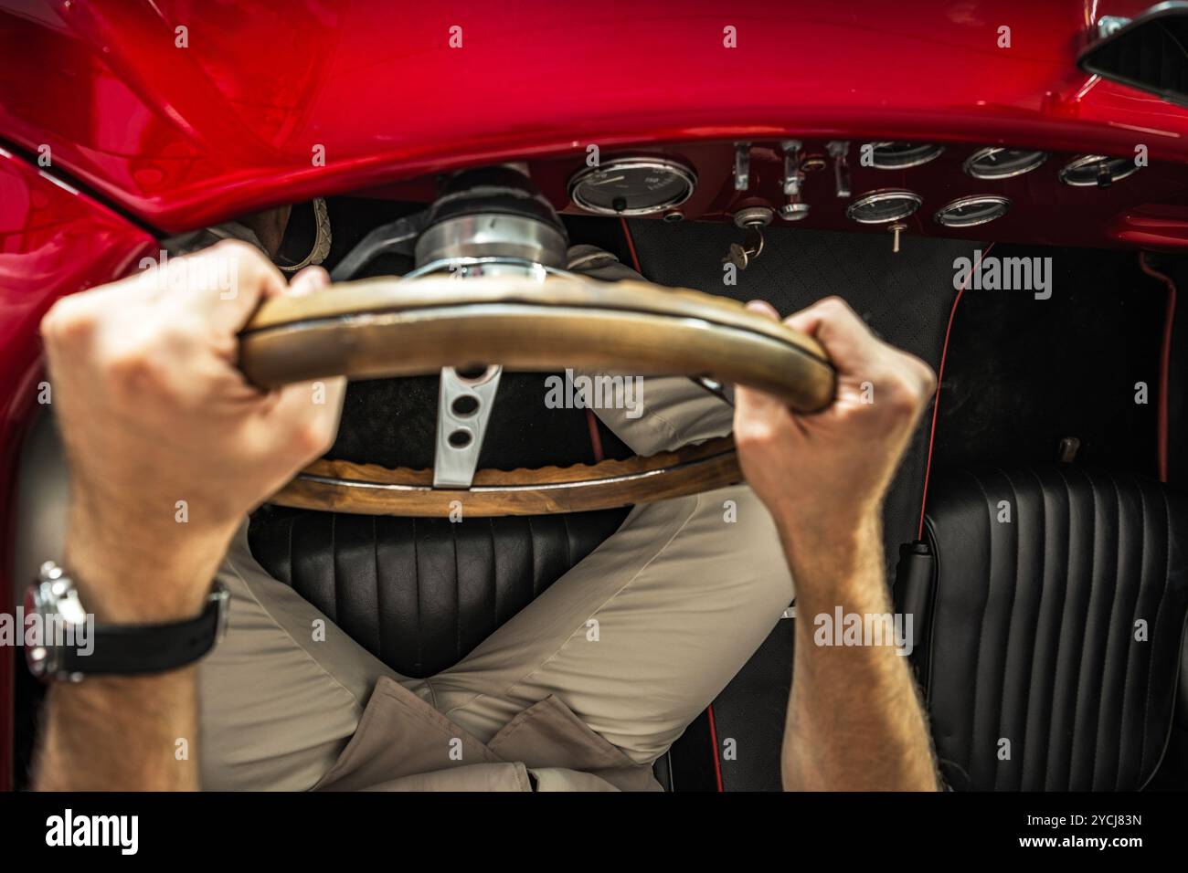 Eine Person greift das hölzerne Lenkrad eines klassischen roten Autos und konzentriert sich auf das Fahren auf einer offenen Straße unter hellem Sonnenlicht. Stockfoto