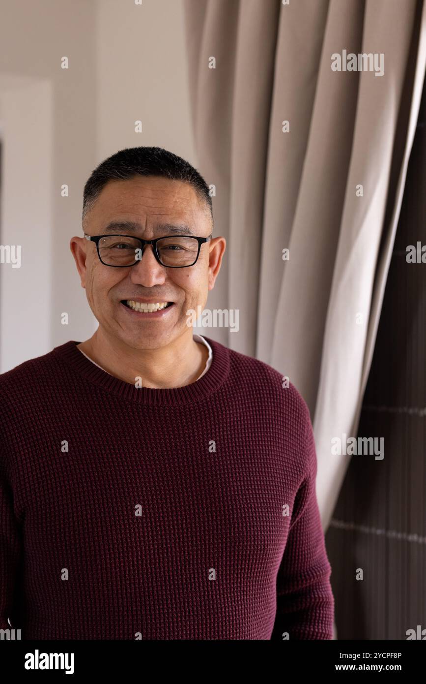 Lächelnder asiatischer Seniorenmann mit Brille und Pullover, der zu Hause neben dem Fenster steht Stockfoto