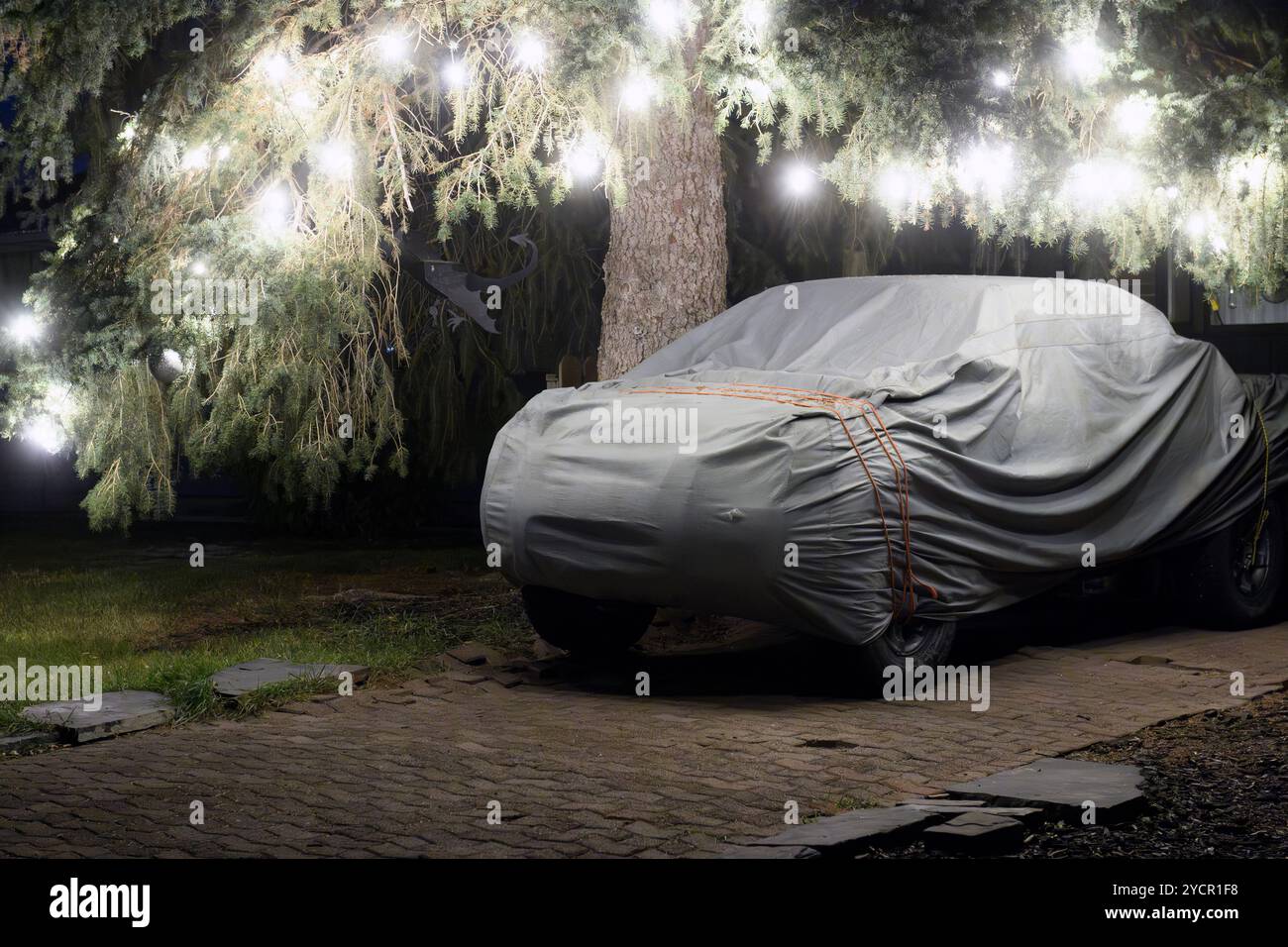 Auto bedeckt mit einer Plane unter einem Baum, dekoriert mit leuchtenden Lichtern bei Nacht, in einer Vorstadteinfahrt Stockfoto