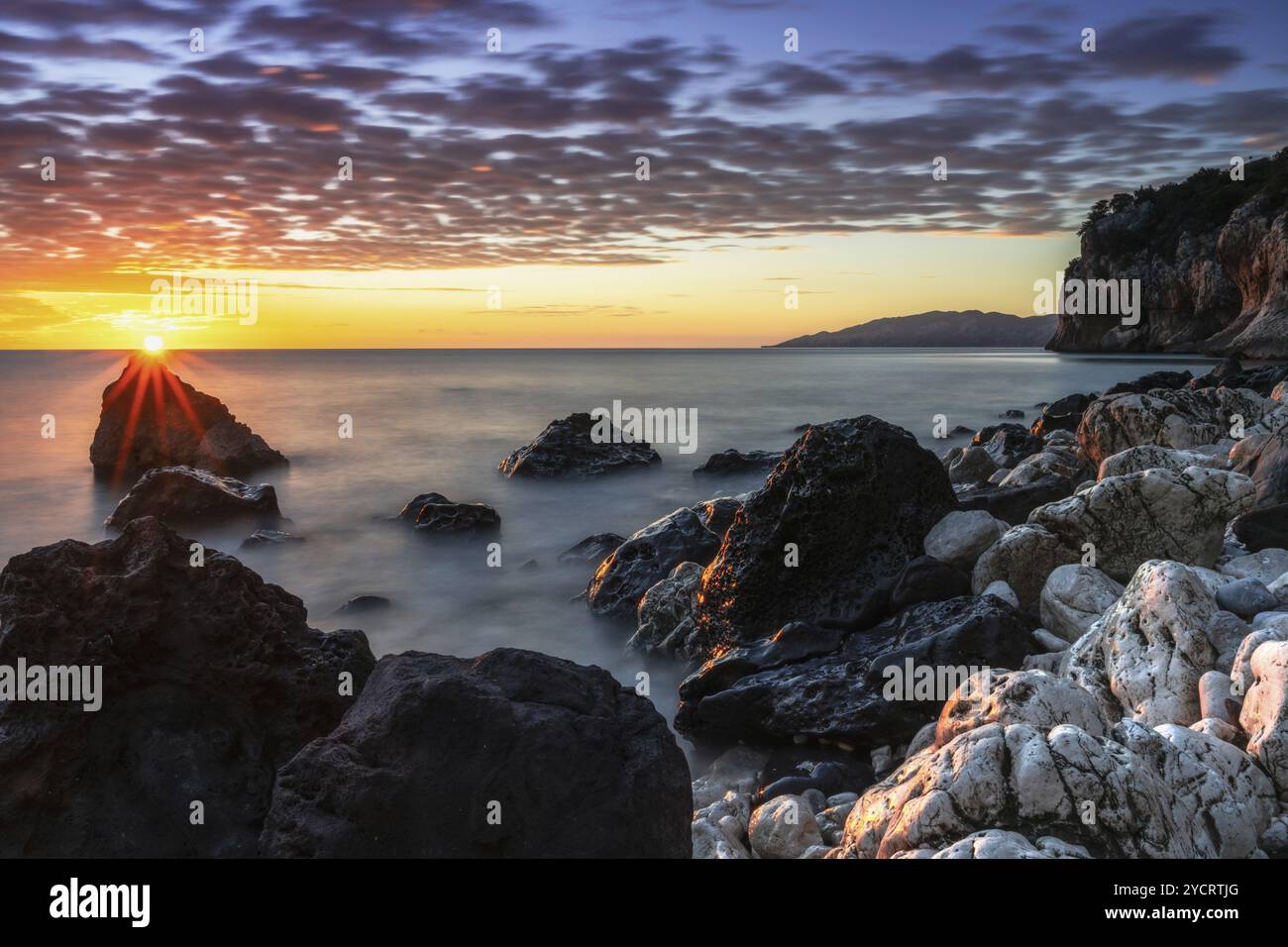 Ein farbenfroher Sonnenaufgang in Cala Gonone mit schwarz-weißen Felsen und Felsbrocken im Vordergrund Stockfoto