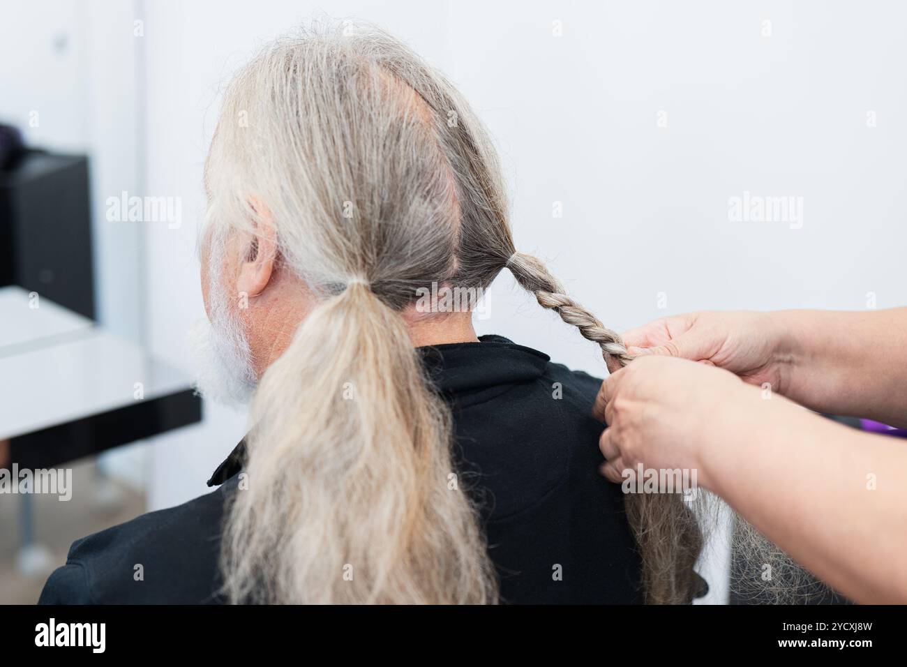 Ein älterer Mann mit langen grauen Haaren bekommt seinen Pferdeschwanz in einem lokalen Friseursalon zur Spende geflochten, was zu einer wohltätigen Sache beiträgt Stockfoto