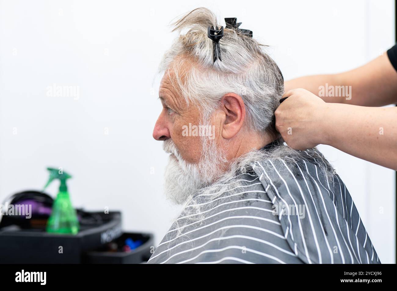 Ein älterer Mann mit grauem und weißem Haar sitzt in einem Friseurstuhl, mit abgeschnittenen und gefesselten Haaren, bereit nach dem Schneiden von Haaren donati Stockfoto