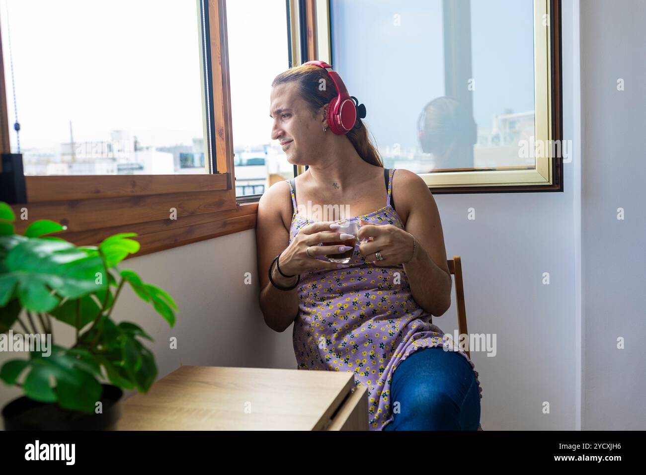 Eine entspannte Transsexuelle hört Musik in einer gemütlichen Umgebung, hält eine weiße Tasse in der Hand und spiegelt die Gelassenheit ihrer morgendlichen Routine wider Stockfoto