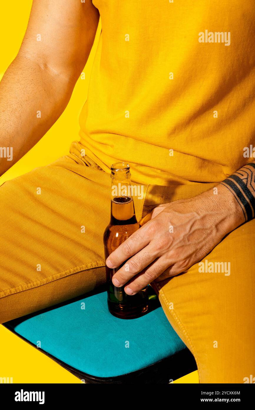 Bei einer Nahaufnahme wird eine anonyme Person in leuchtend gelber Kleidung gefangen, die eine Bierflasche vor einem kräftigen gelben Hintergrund hält Stockfoto