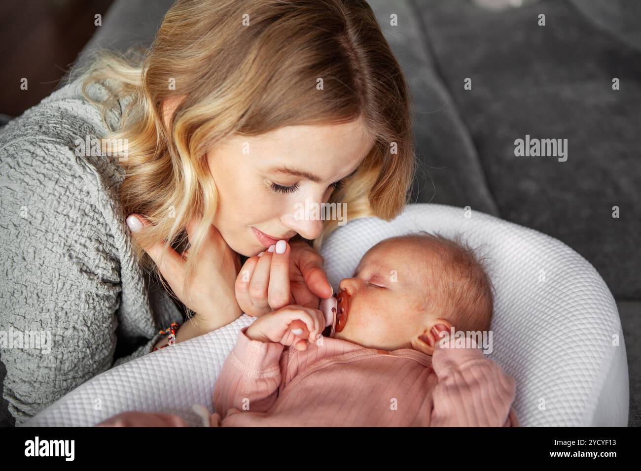 Die schöne Mutter blickt mit Liebe auf das schlafende Neugeborene, das in rosa Einteiler gekleidet ist und einen Schnuller im Mund hat. Ein ruhiger Moment zu Hause, spiegelt tiefe Verbundenheit wider Stockfoto