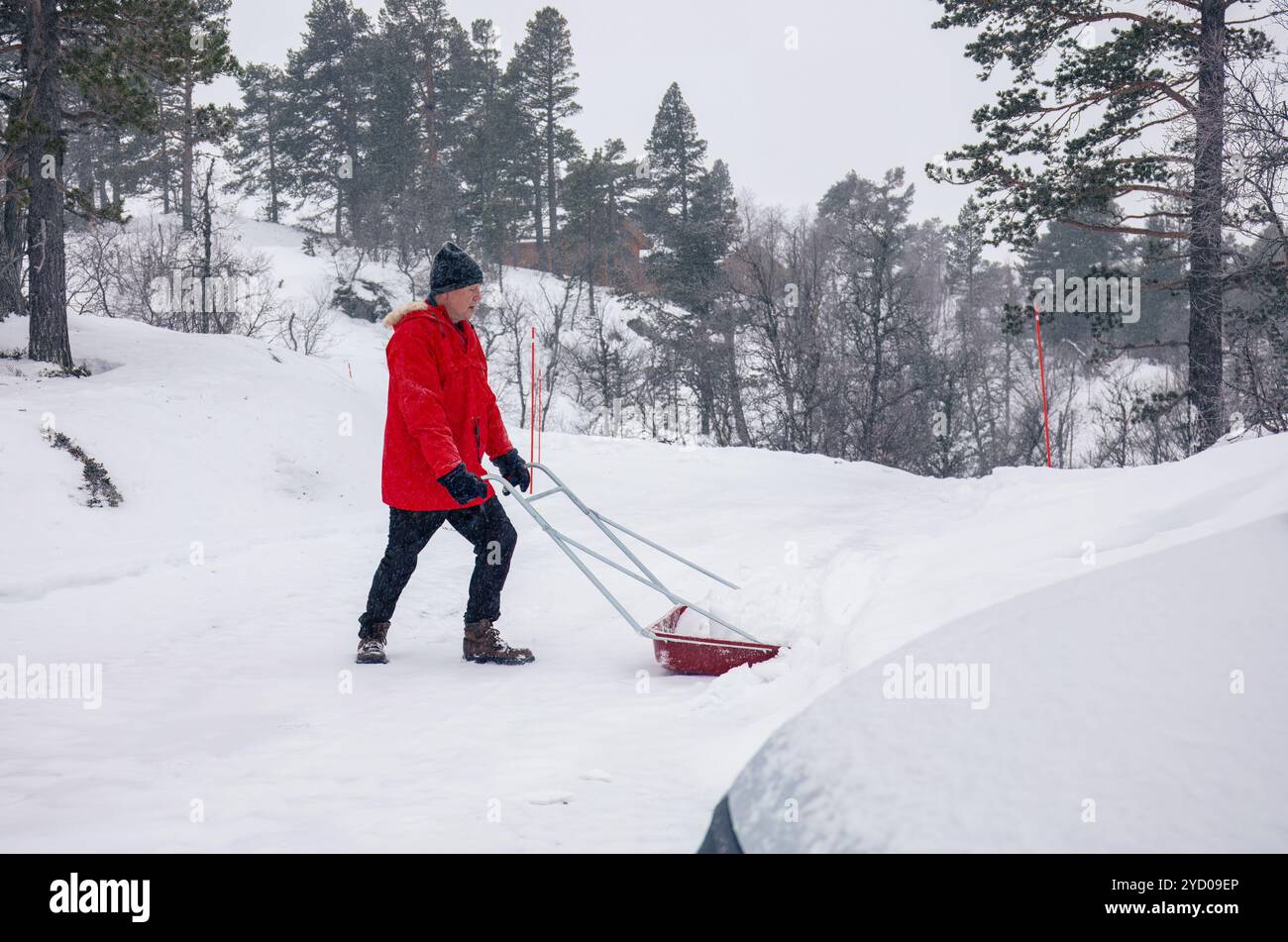 Mann in roter Jacke schiebt einen Schneeschieber auf einer schneebedeckten Straße durch Berghütte, Schneeräumung, Winterarbeiten, ländliches Leben, raues Wetter Stockfoto