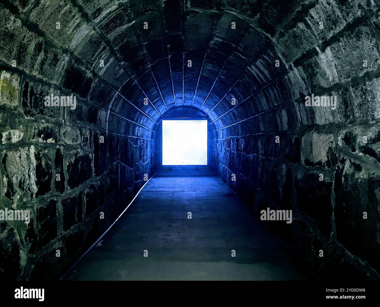 Ein schwach beleuchteter Steintunnel mit einem geheimnisvollen blauen Leuchten am Ende, der ein schauriges Halloween-Ambiente schafft Stockfoto