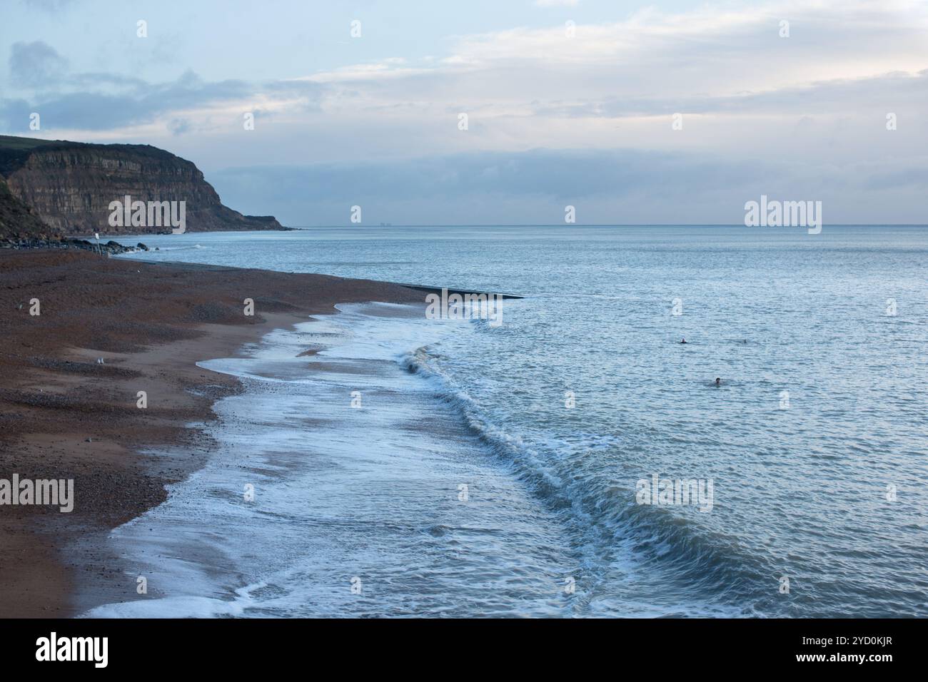 Ruhige Küstenlinie am Rock-a-Nore Beach in Hastings, mit ruhigen Wellen und weit entfernten Winterschwimmern unter bewölktem Himmel. Stockfoto