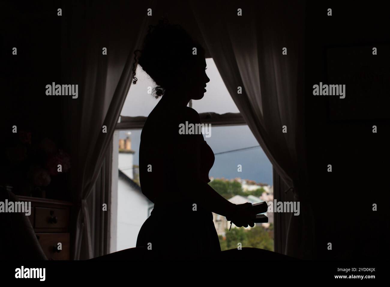 Silhouette einer Frau an einem Fenster mit Vorhängen, das ein Objekt hält, vor einem weichen, natürlichen Hintergrund und einem bewölkten Himmel. Stockfoto