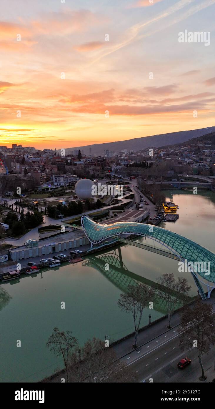 Die Friedensbrücke, die bei Sonnenuntergang in Tiflis, Georgien, beleuchtet wird, überspannt den Fluss Kura mit dramatischem orange-rosa Himmel über der Hauptstadt. Stockfoto