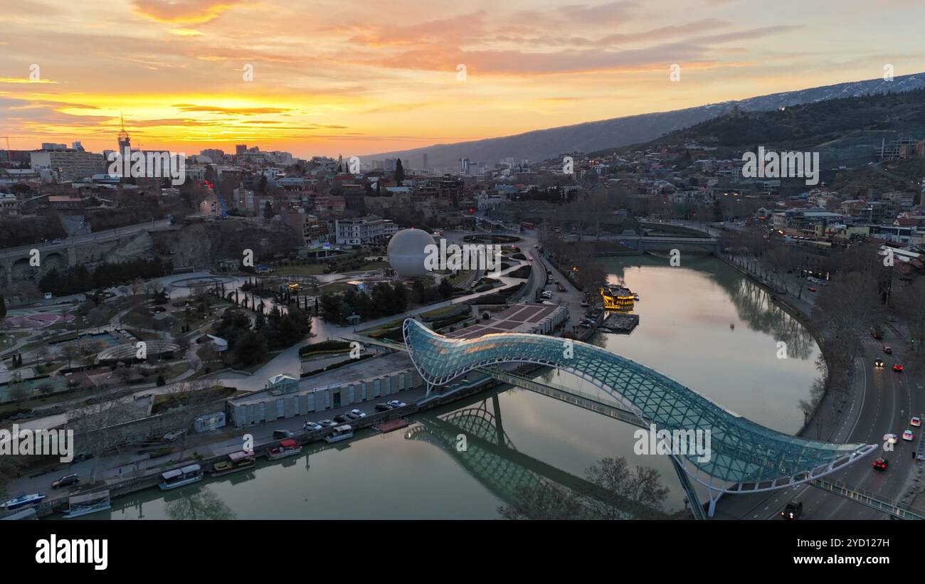 Die ikonische Friedensbrücke überspannt den Fluss Kura bei Sonnenuntergang in Tiflis, Georgien, mit einer Glas- und Stahlkonstruktion, die vor einem goldenen Himmel beleuchtet wird. Stockfoto