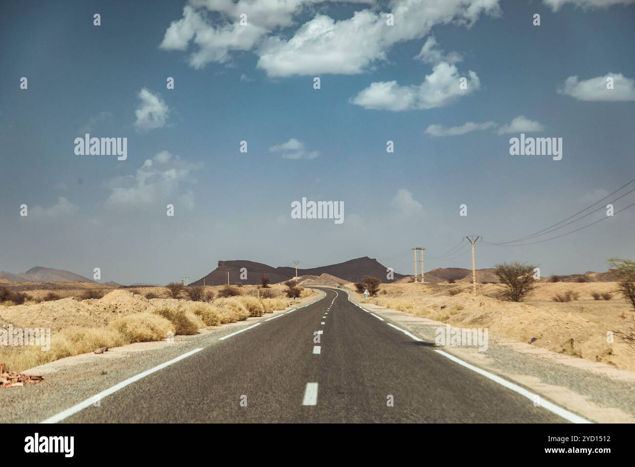 Ein langer Abschnitt der Autobahn verschwindet in Marokko am Horizont, umgeben von trockenem Grasland und fernen Hügeln unter Cumulus-Wolken. Die helle Sonne Stockfoto