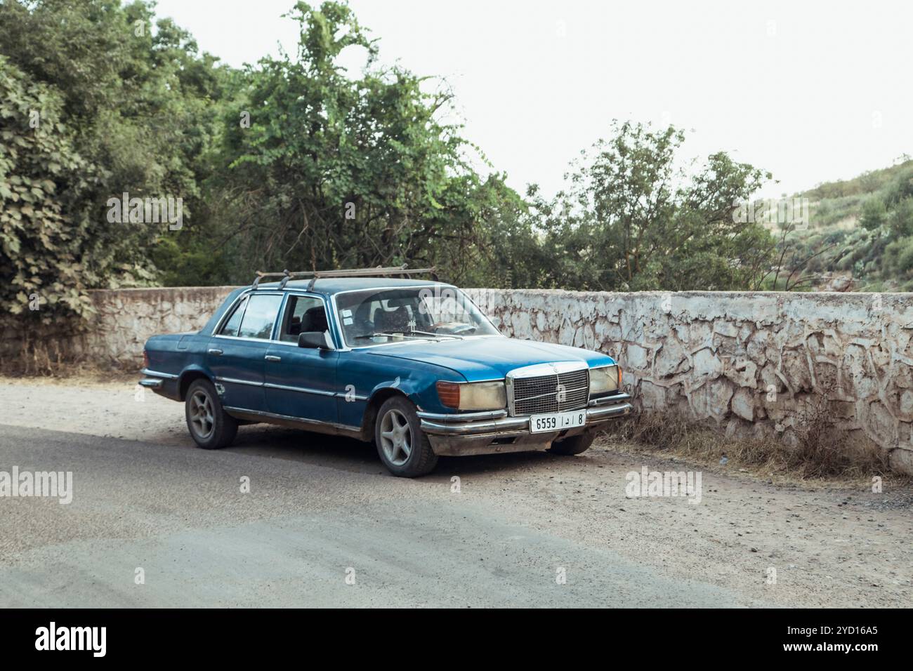 Ein Oldtimer-Luxusauto parkt neben einer Steinmauer in Marokko, umgeben von Grün und einer ruhigen Atmosphäre. Das Fahrzeug zeigt sein klassisches Design Stockfoto