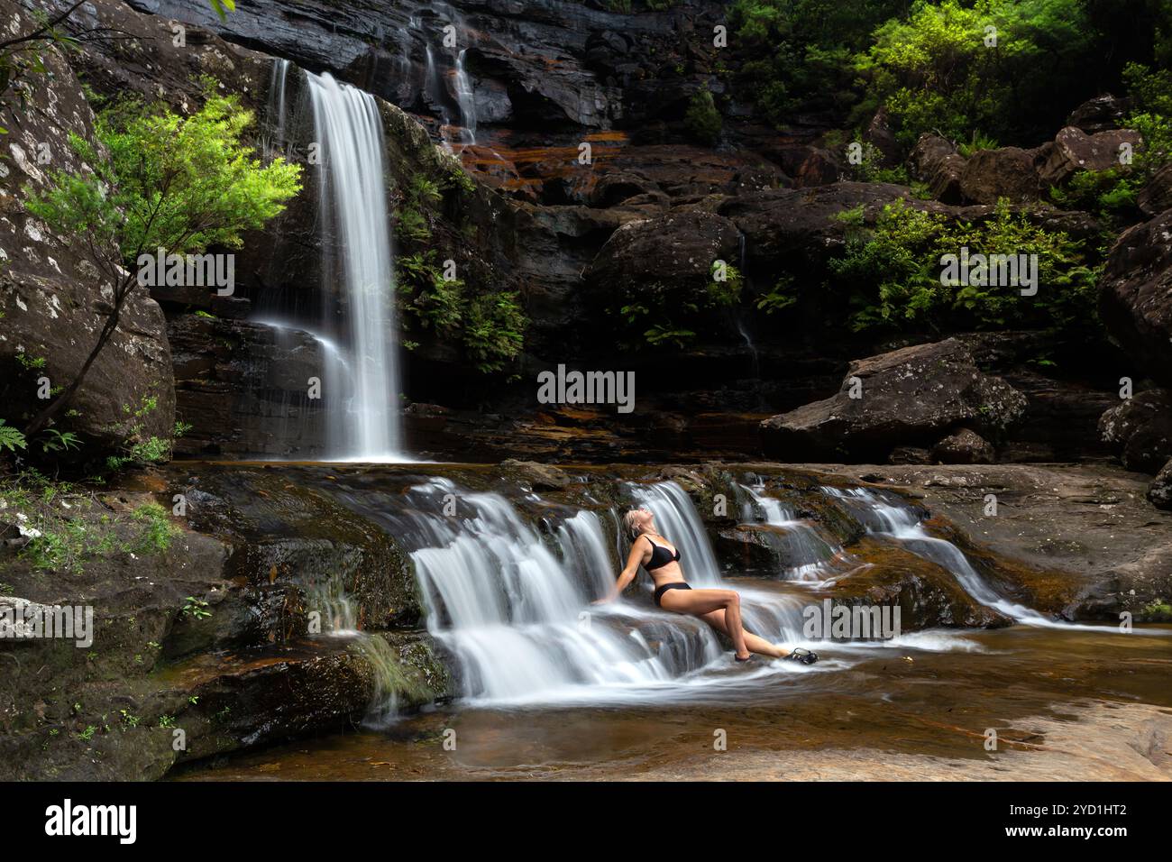 Frau, die in fließenden Wasserfallkaskaden sitzt und in einer Naturoase eingetaucht ist Stockfoto